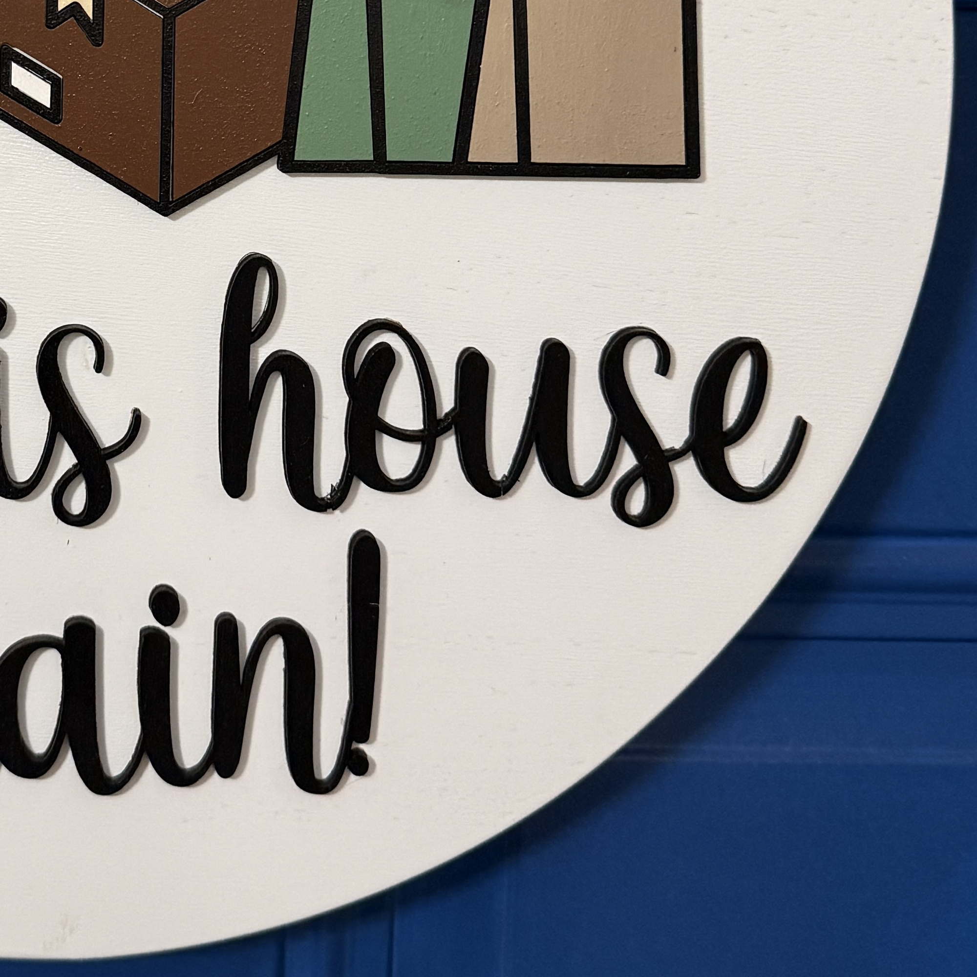 Close-up of raised black script text reading “Yes, this house again!” on round white wooden door hanger against blue door background.