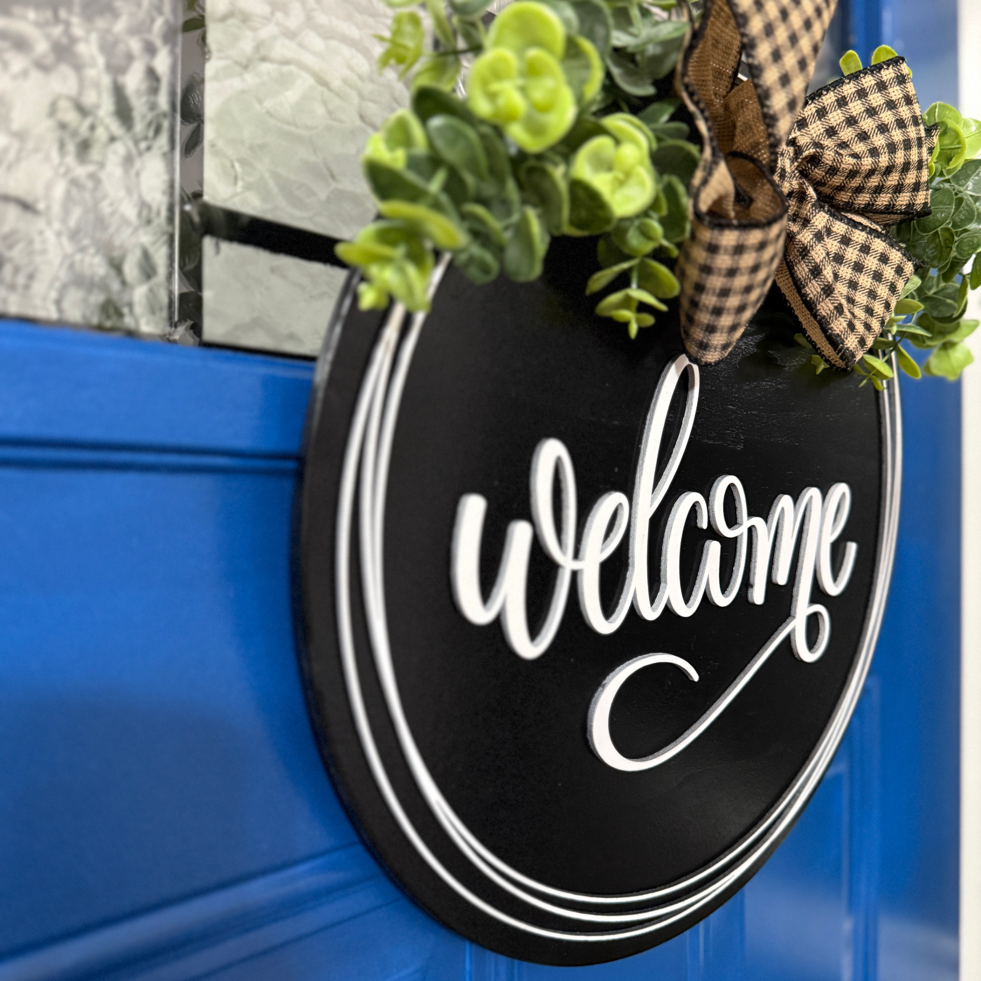 Angled view of black welcome door hanger with layered white border detail, greenery, and plaid ribbon bow