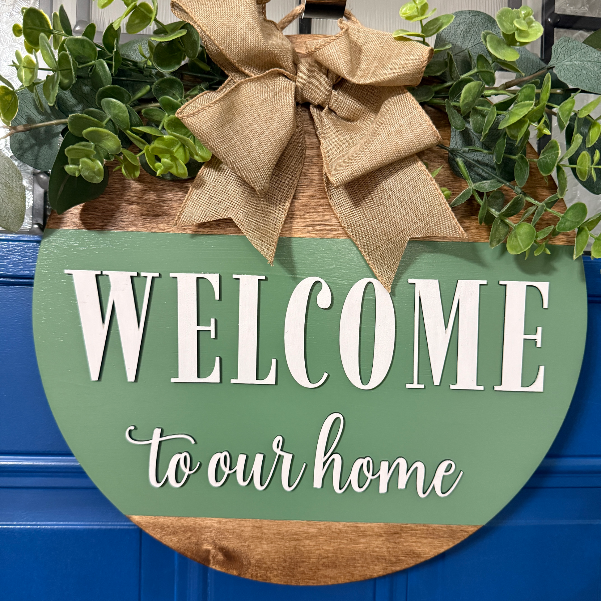 Sage green welcome to our home door hanger with greenery accents and burlap bow hanging on blue front door
