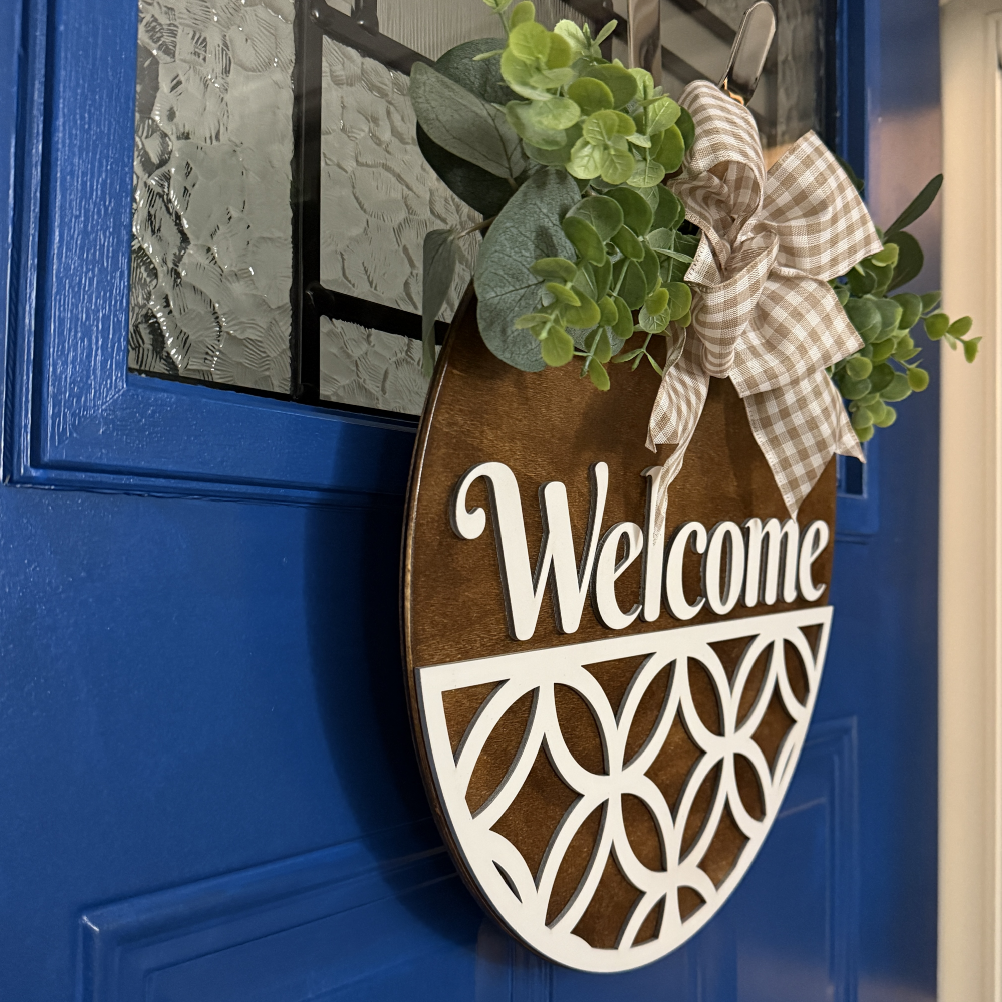 Side view of rustic welcome door hanger with white geometric pattern, gingham bow, and greenery accents