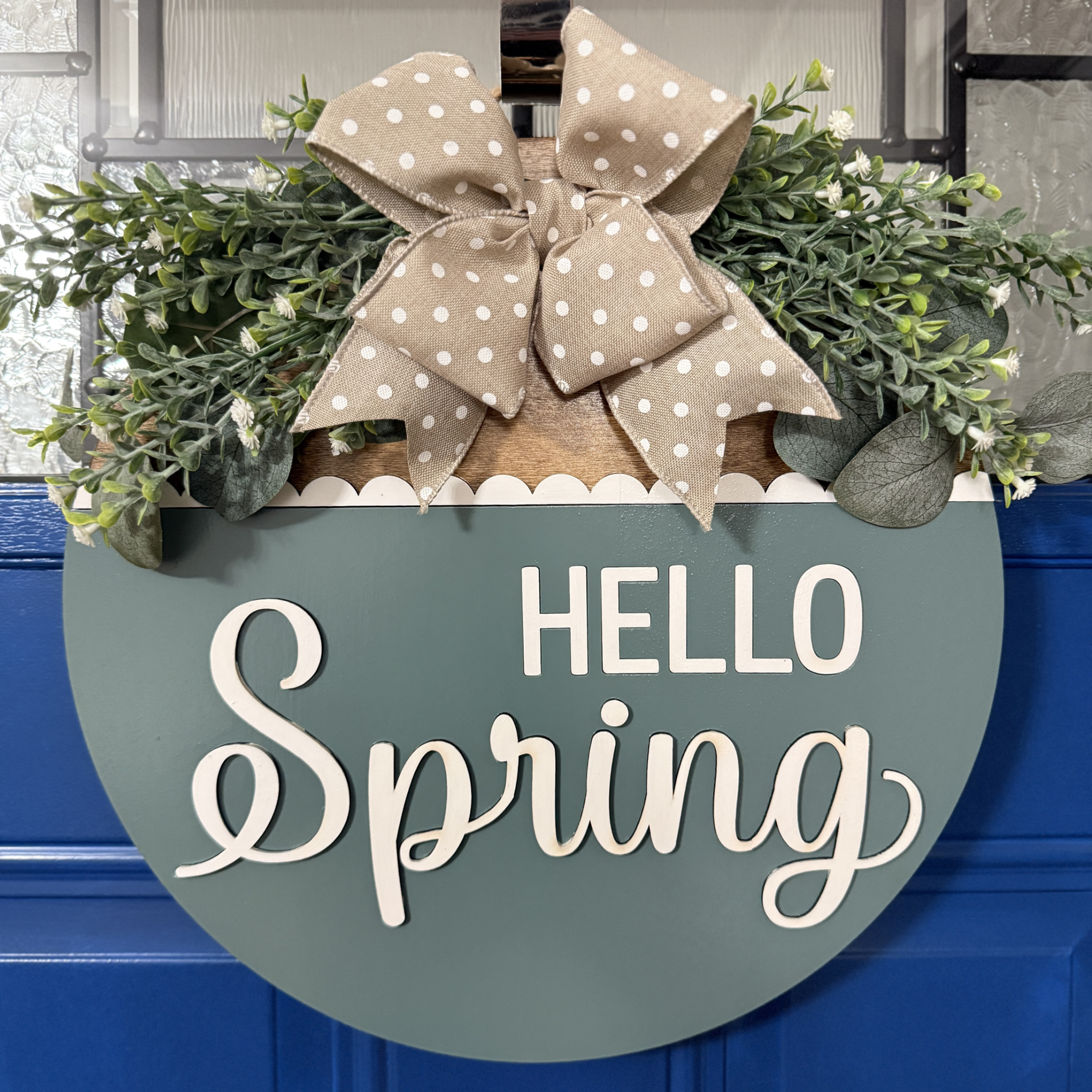 Front view of patina blue Hello Spring door hanger with white raised lettering, layered greenery, and neutral polka dot bow against a blue front door