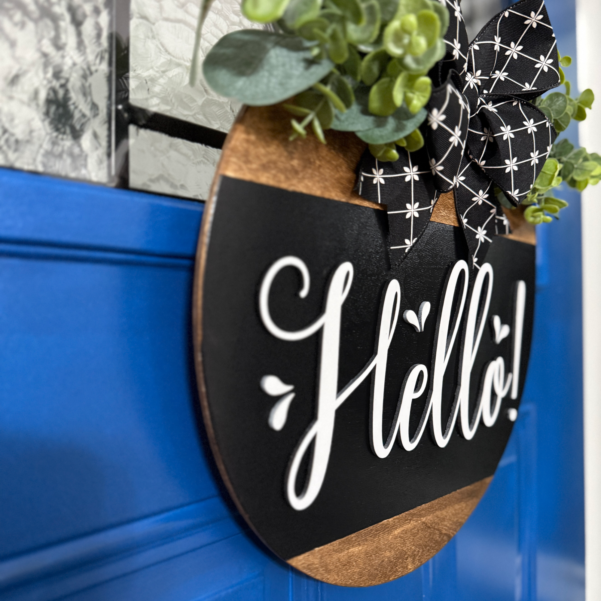 Angled view of black Hello door hanger with eucalyptus greenery and black ribbon bow hanging on blue door