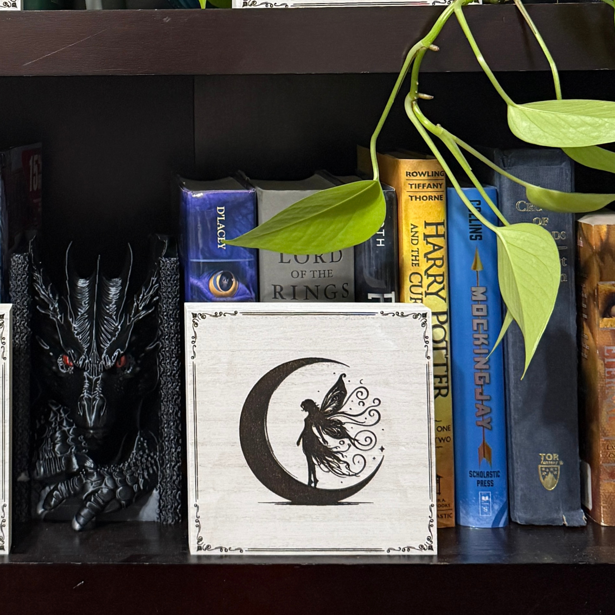 Moon fairy shelf sitter on a bookshelf beside fantasy novels and dragon decor, highlighting engraved crescent moon fairy artwork