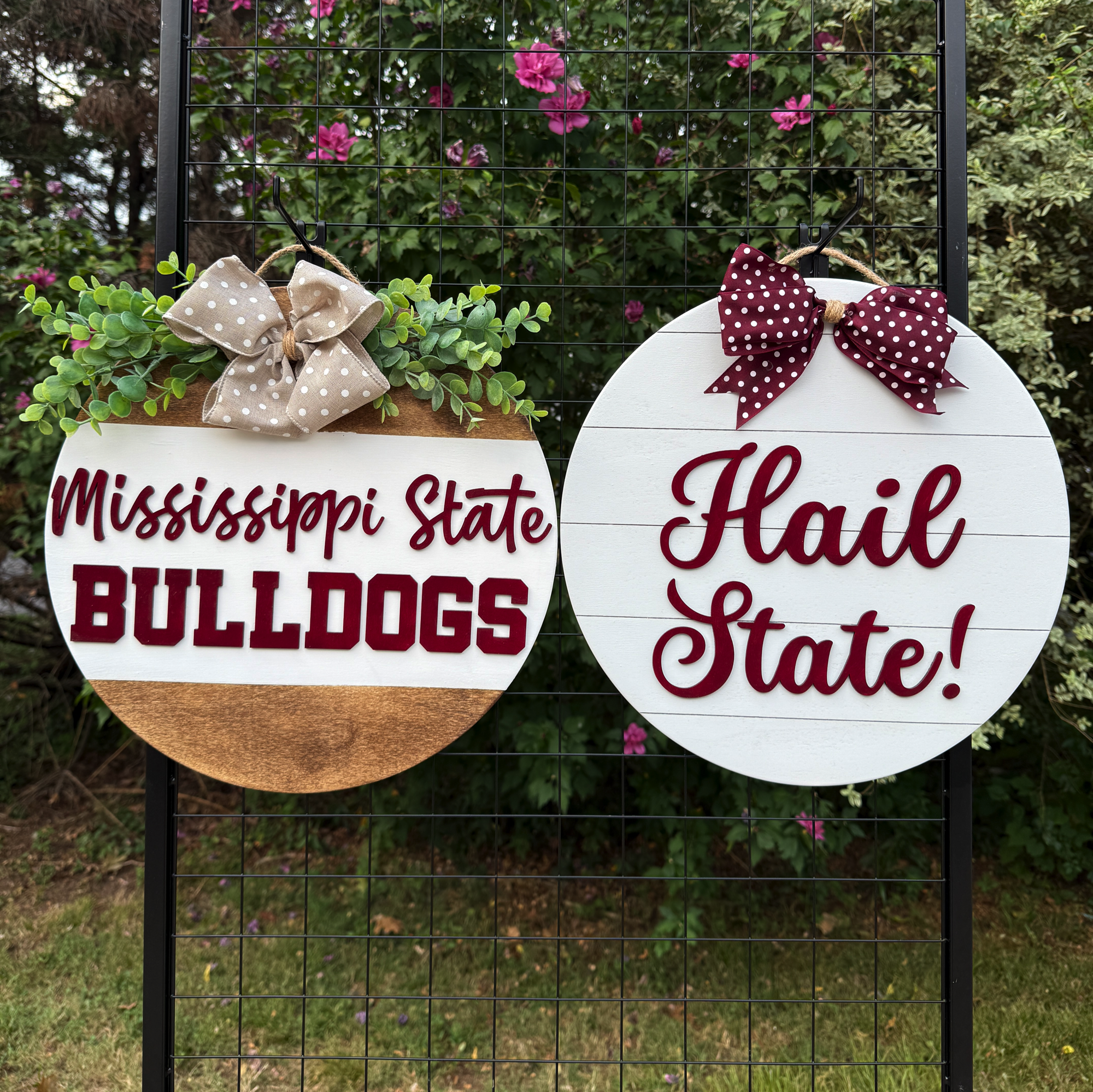 Two round Mississippi State-themed door hangers displayed outdoors on a black grid with greenery in the background; one reads “Mississippi State Bulldogs” and the other “Hail State!”