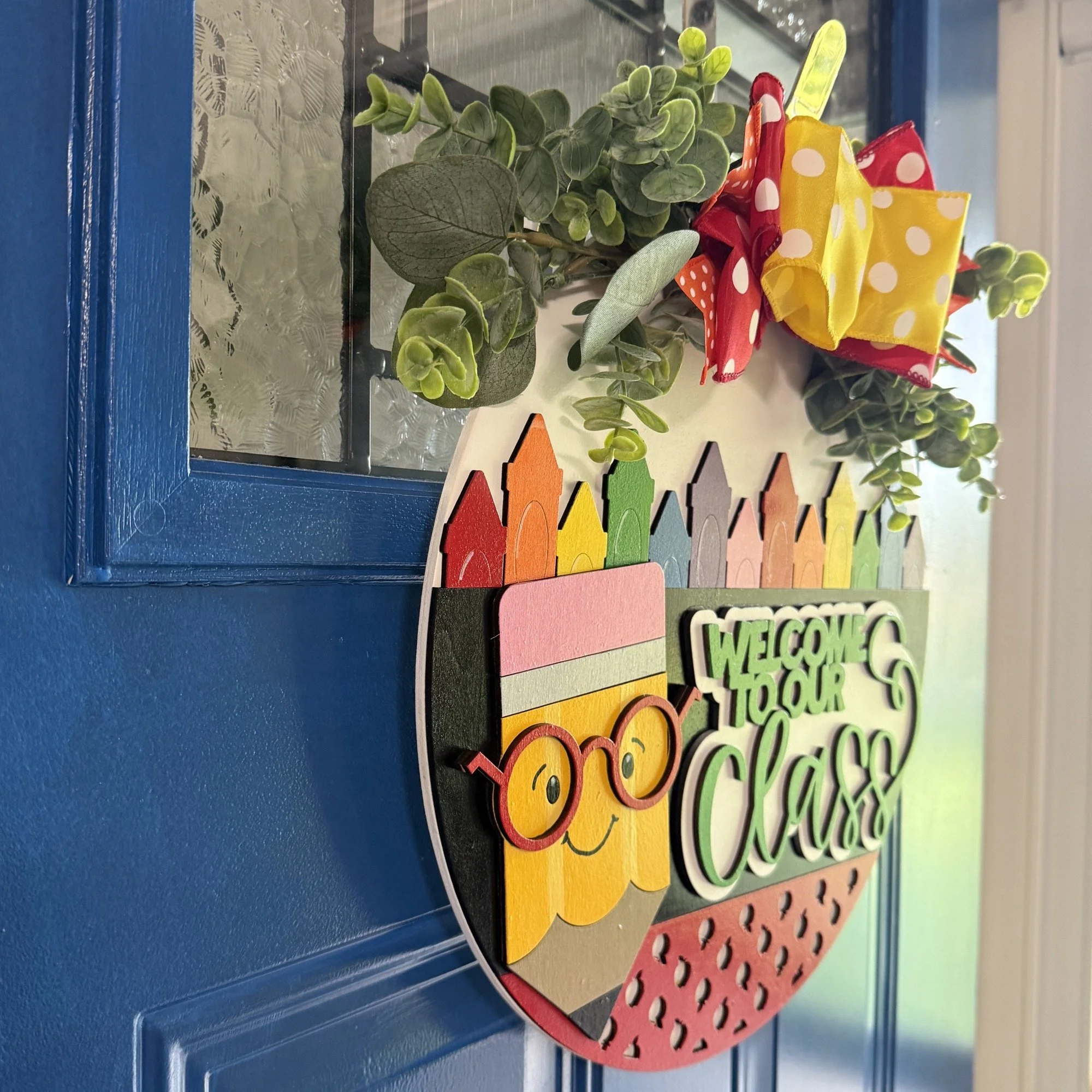 Side view of a colorful teacher-themed door hanger with rainbow pencil border, red polka dot apples, “Welcome to Our Class” text, and layered bow with greenery on top