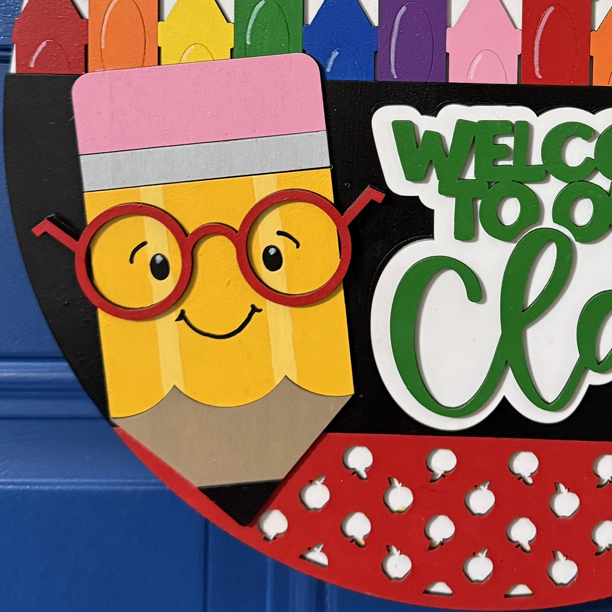 Side view of “Welcome to Our Class” door hanger featuring smiling pencil, rainbow crayon row, and red/yellow polka dot bow with greenery on a dark blue door.