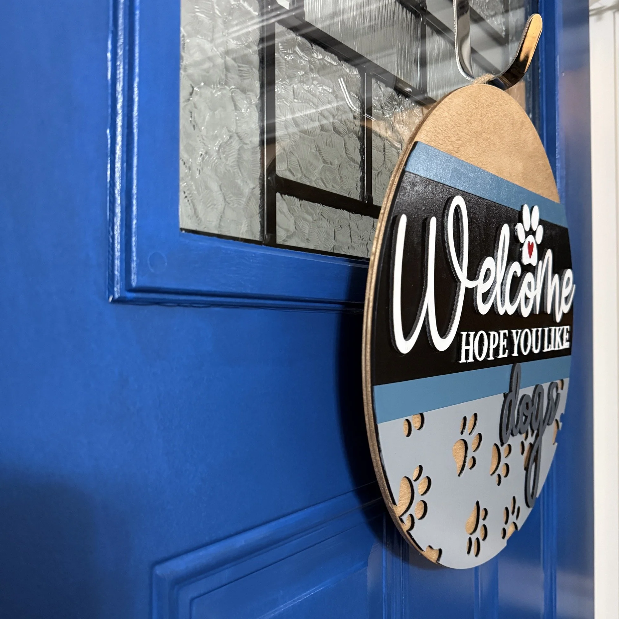 Side view of “Welcome Hope You Like Dogs” door hanger with paw prints, heart accent, and neutral colors, hanging on a blue front door.