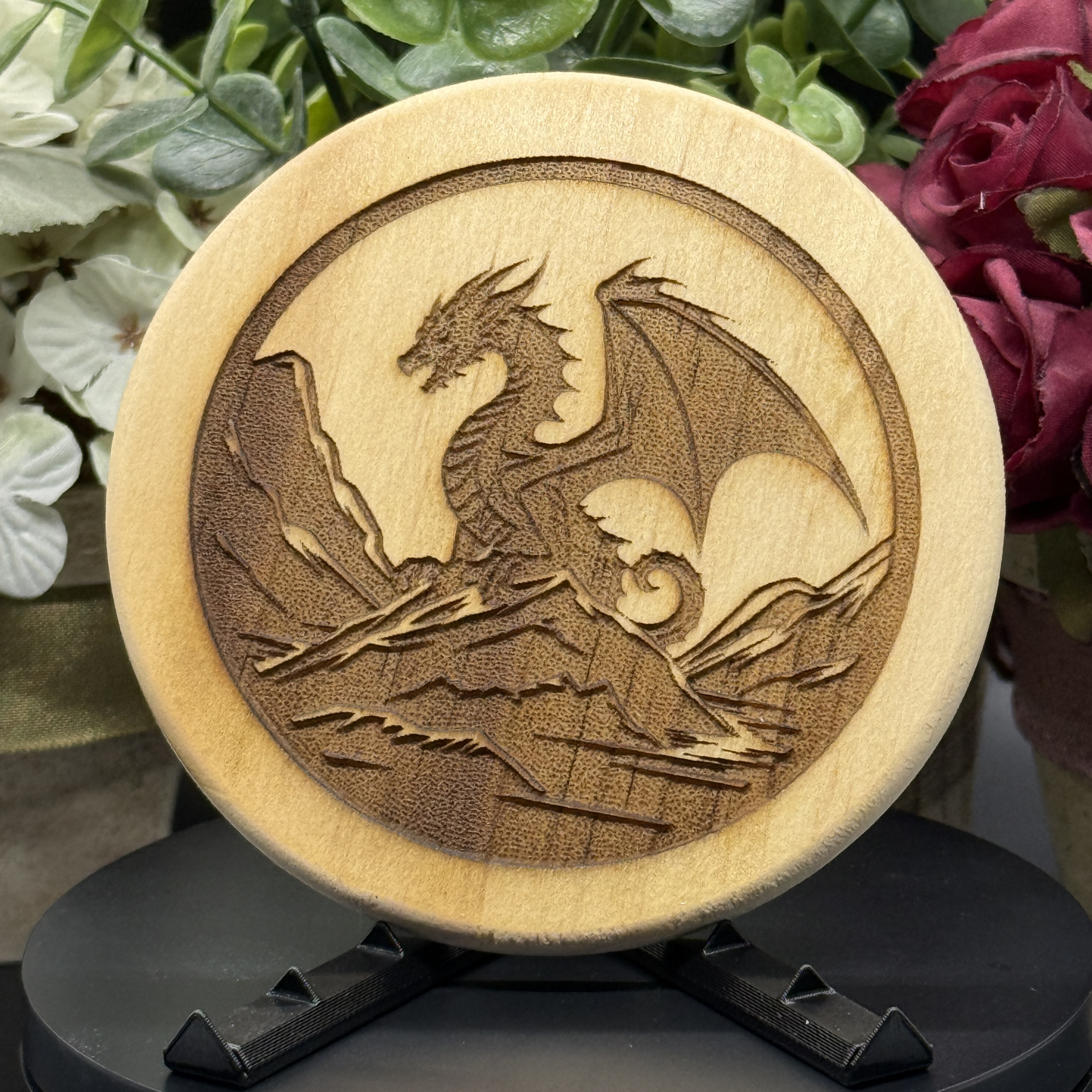 Round pine wood coaster with dragon silhouette perched above waves or mountains, tail curled and wings raised