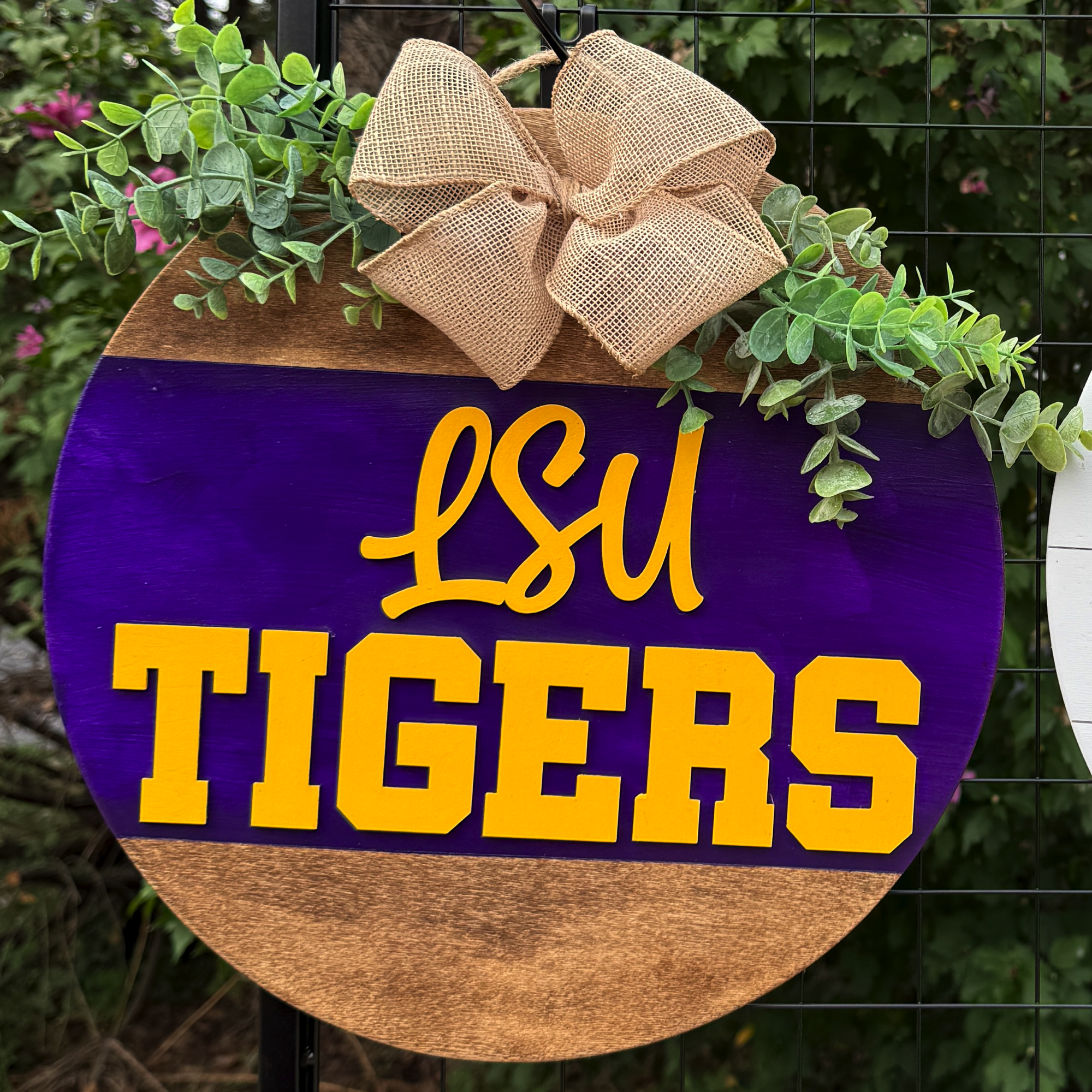 Round LSU Tigers door hanger with purple and gold lettering, burlap bow, and greenery accents on rustic wood background