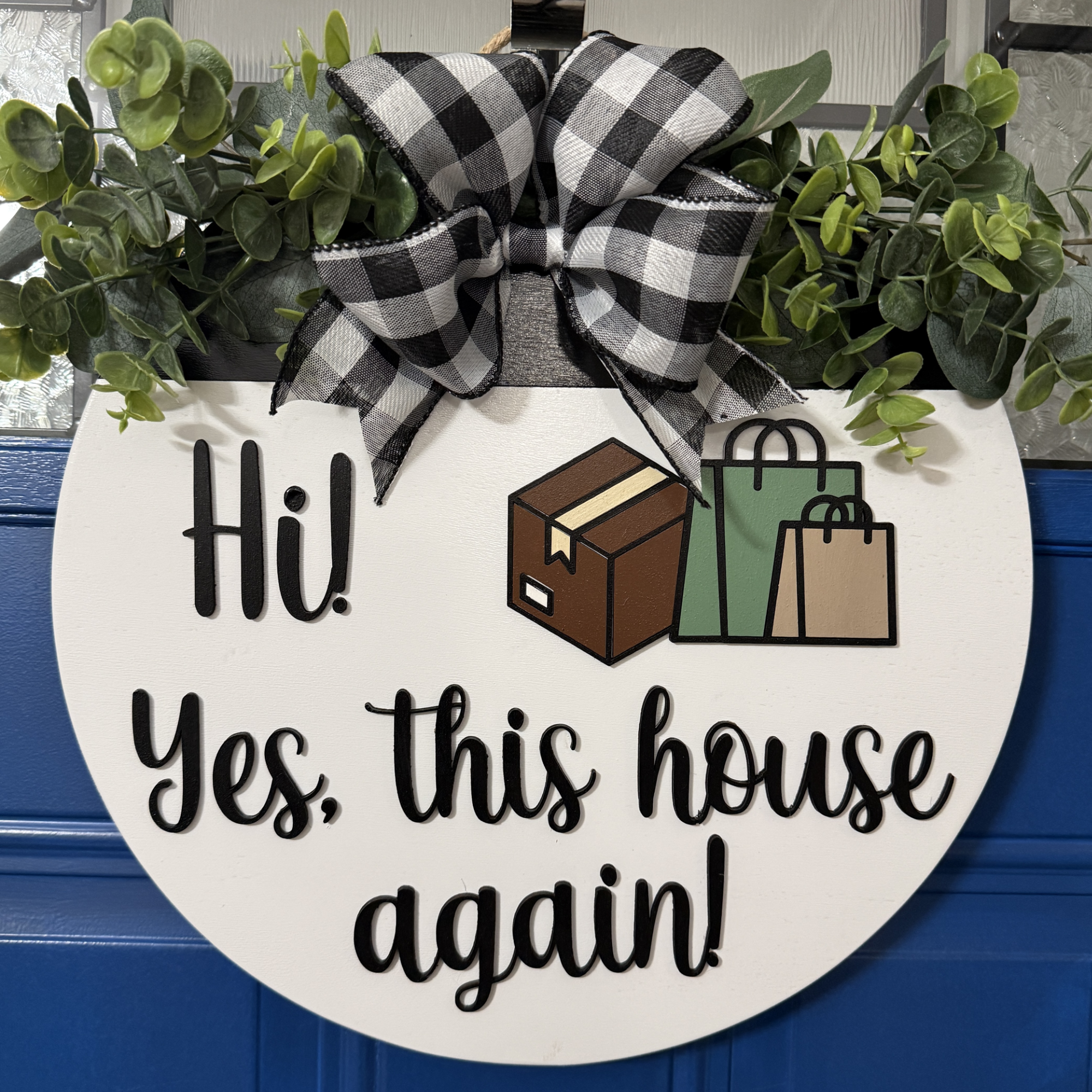 Front view of round white wooden door hanger reading “Hi! Yes, This House Again” with layered package and shopping bag graphics, black and white buffalo plaid bow, greenery, displayed on a blue door.