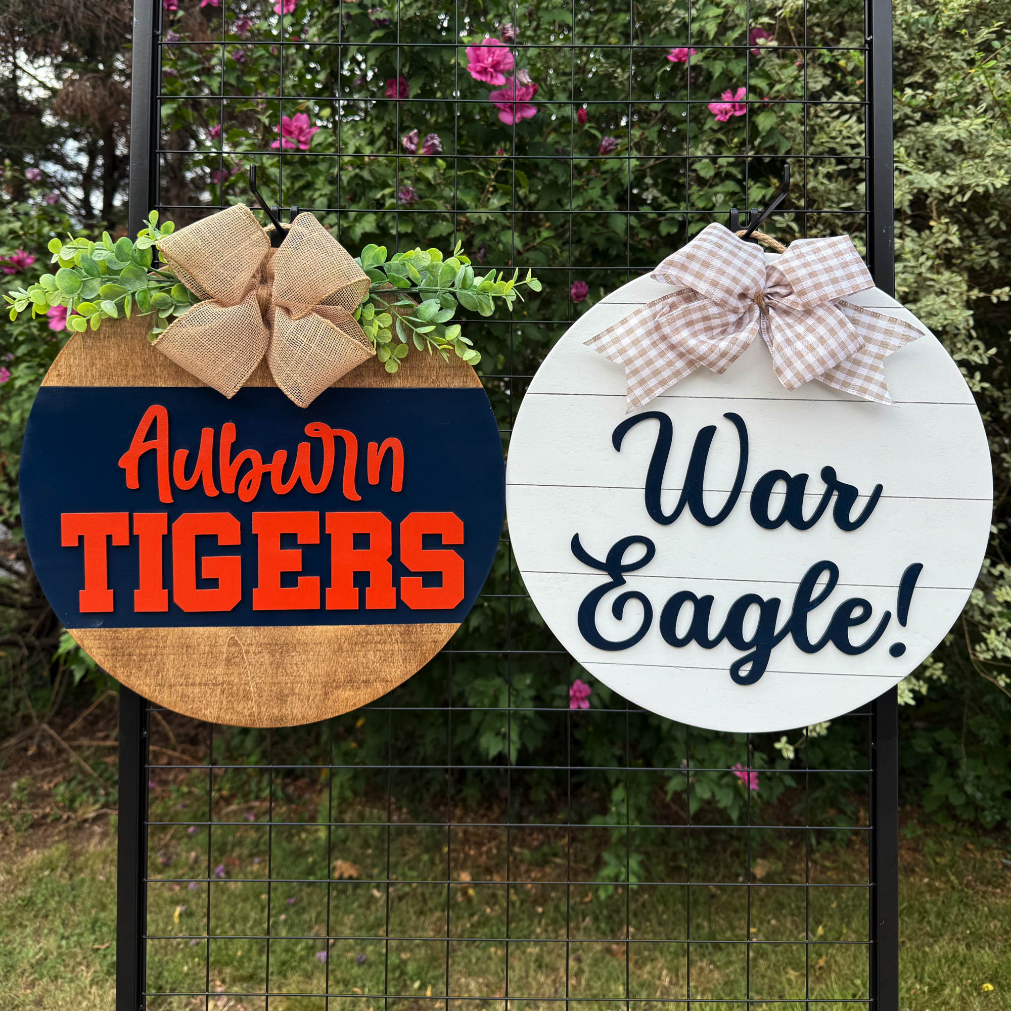 Two round Auburn-themed door hangers displayed outdoors on a black grid with greenery and flowers in the background; one reads “Auburn Tigers” and the other “War Eagle!”
