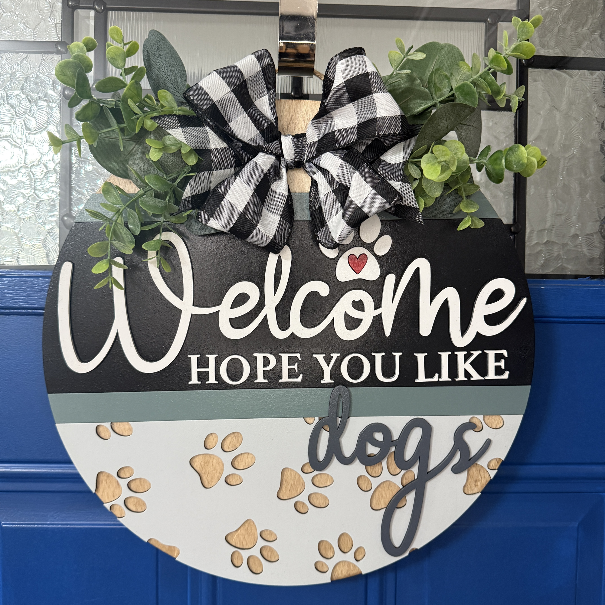 14-inch layered wood “Welcome Hope You Like Dogs” door hanger with paw prints, heart accent, and neutral colors, hanging on a blue front door.