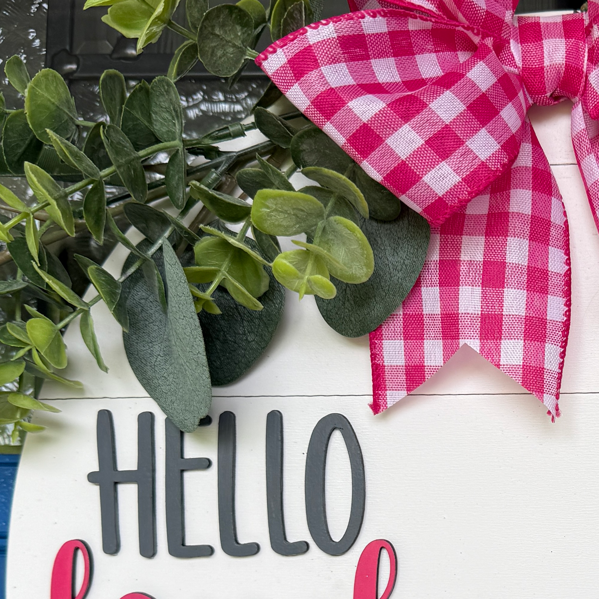 Close-up of pink gingham bow, eucalyptus greenery, and Hello lettering on a handmade watermelon welcome door hanger.