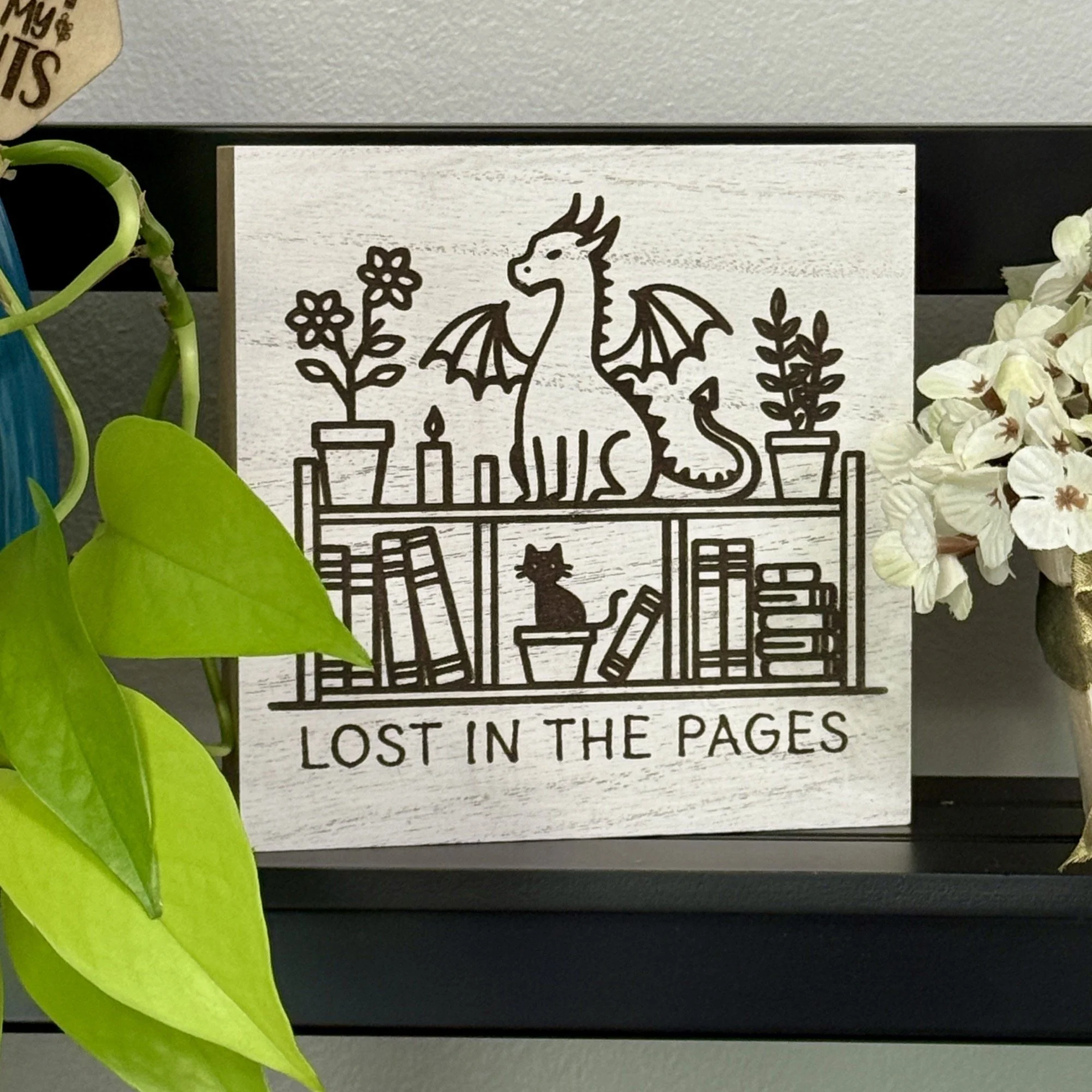 White-washed wood shelf sitter engraved with a seated dragon between potted plants and books, with a small cat figure below, displayed on a black shelf with surrounding greenery and flowers.