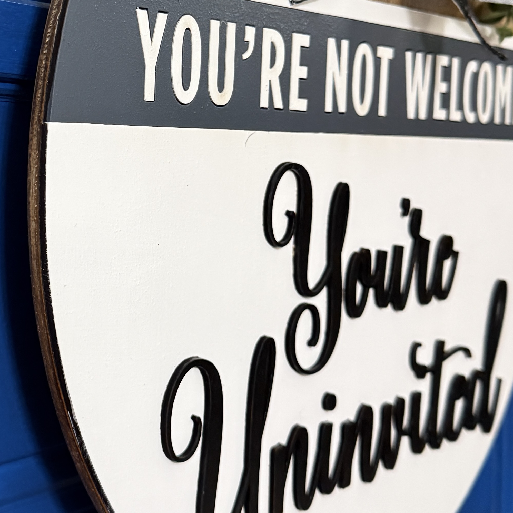 Detail view of dimensional black script lettering along the curved edge of the white wooden door hanger against blue door background.