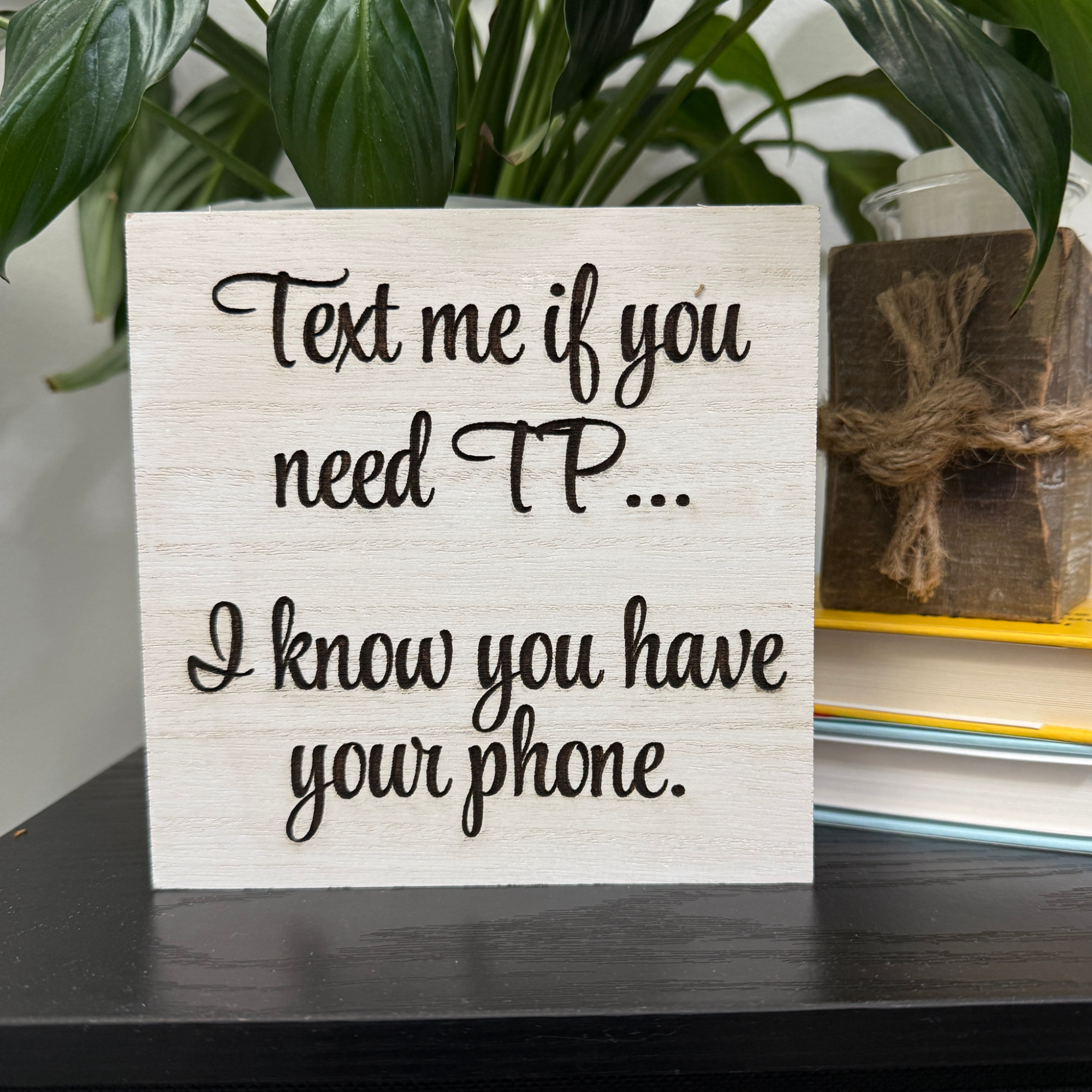 Close-up of humorous bathroom sign with engraved text and smiling poop artwork on white-washed wood, styled next to plant and books