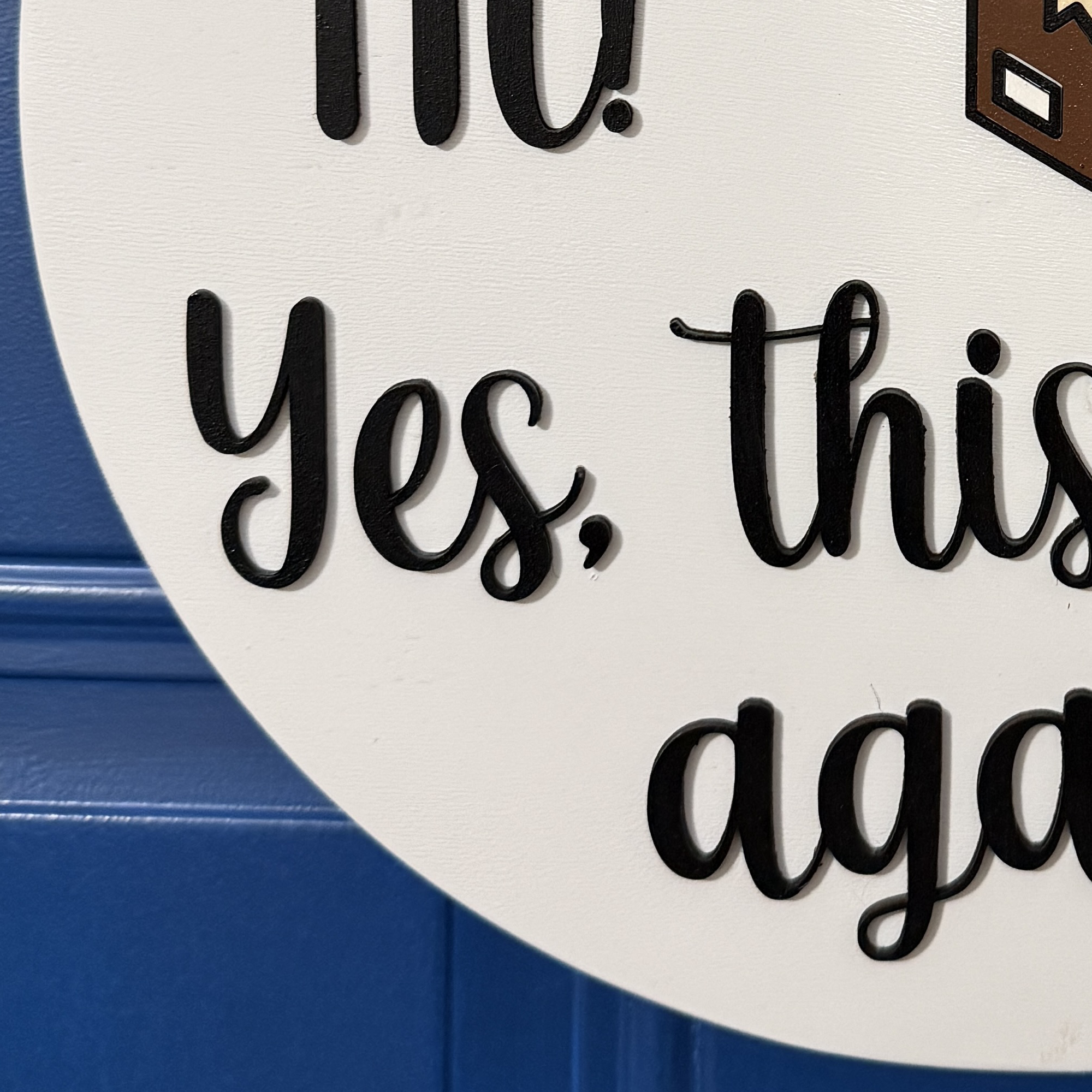 Close-up of raised black script text reading “Yes, this house again!” on round white wooden door hanger against blue door background.