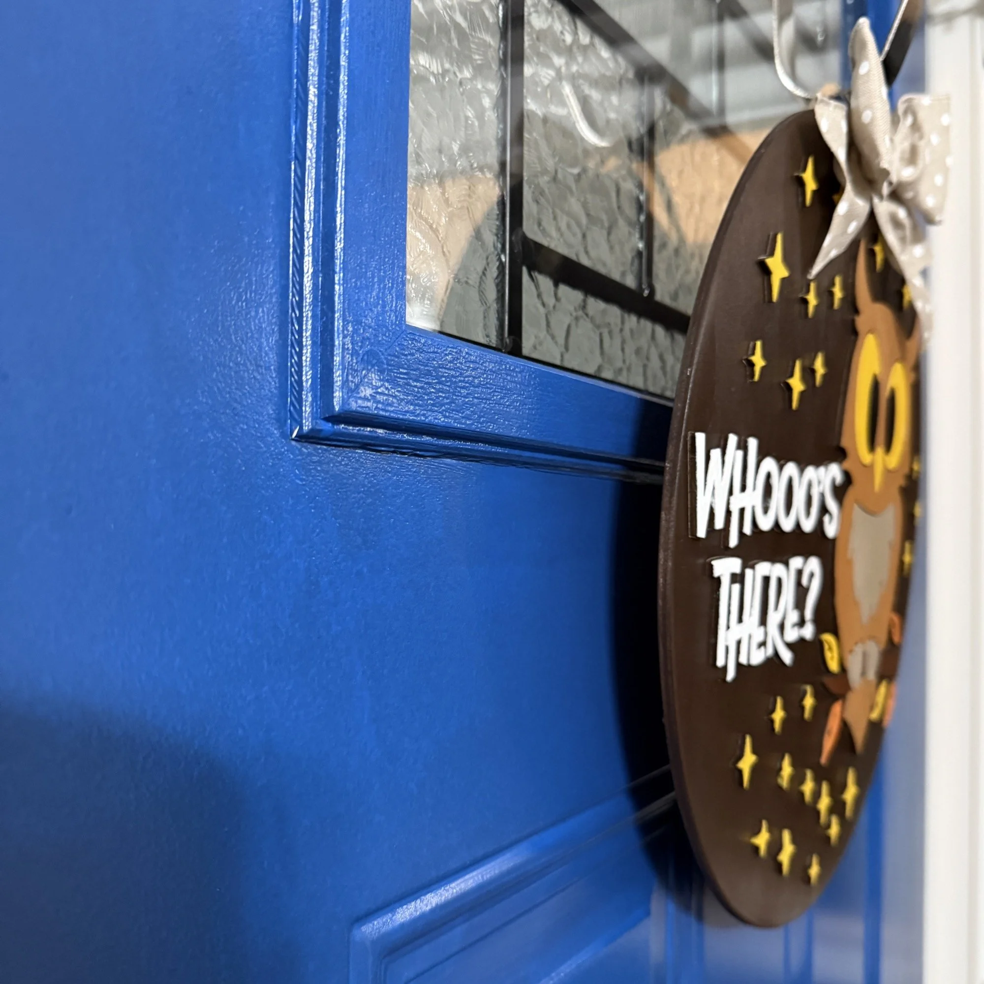 Side view of “Whooo’s There?” fall door hanger with owl, stars, and autumn leaves, topped with a polka dot bow, hanging on a blue front door.