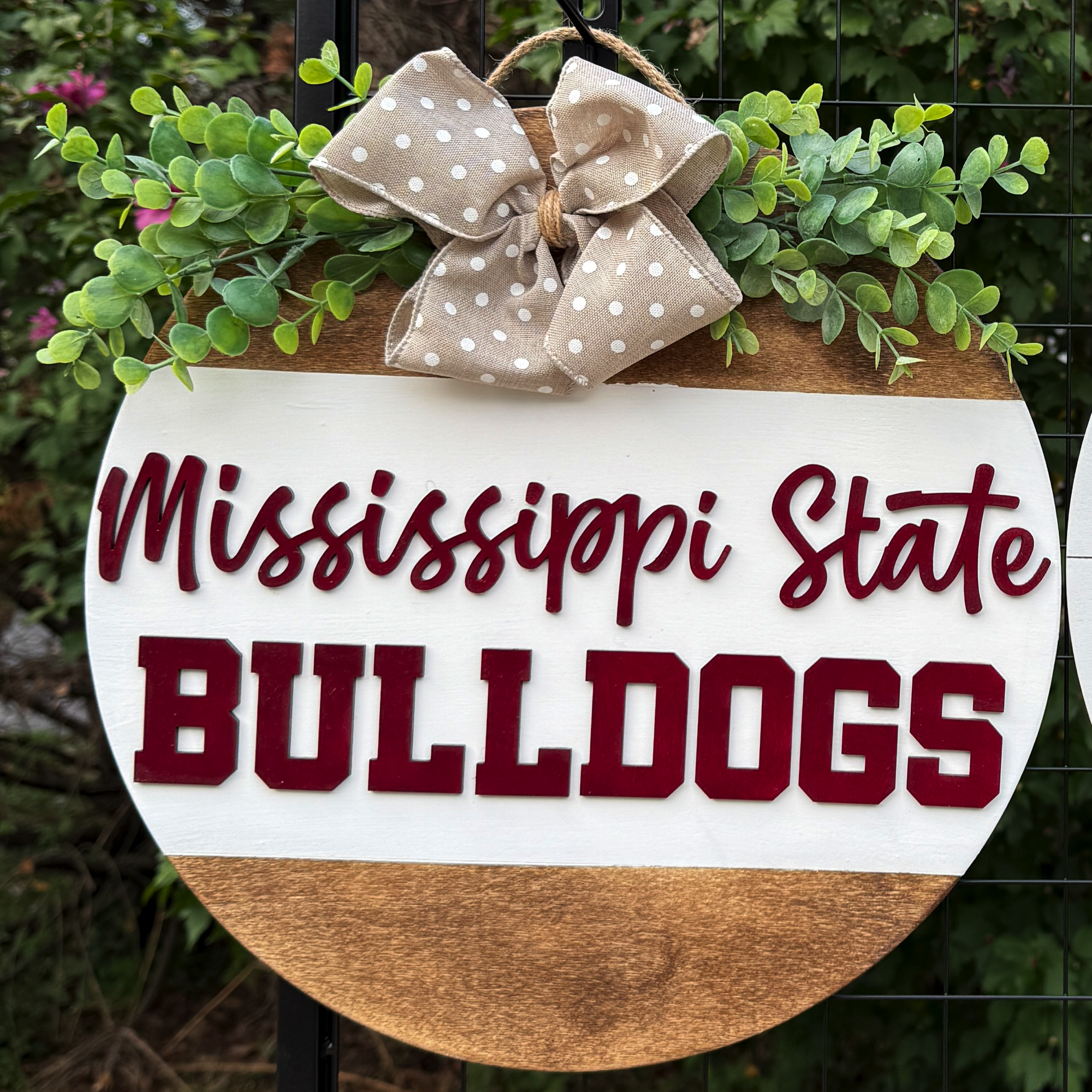 Round Mississippi State Bulldogs door hanger with maroon lettering on white and wood background, greenery accents, and tan polka-dot bow