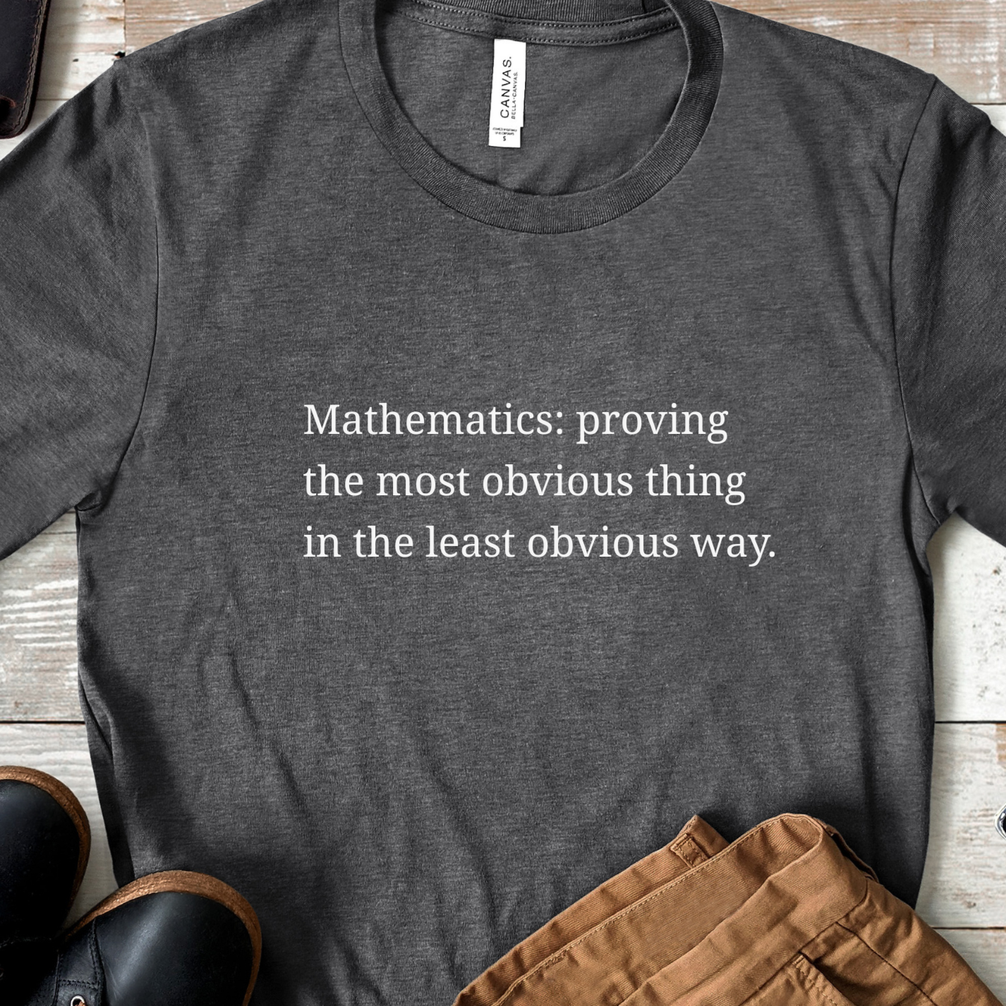 Dark grey heather t-shirt featuring the phrase “Mathematics: proving the most obvious thing in the least obvious way” in soft white text, styled in a casual flat lay