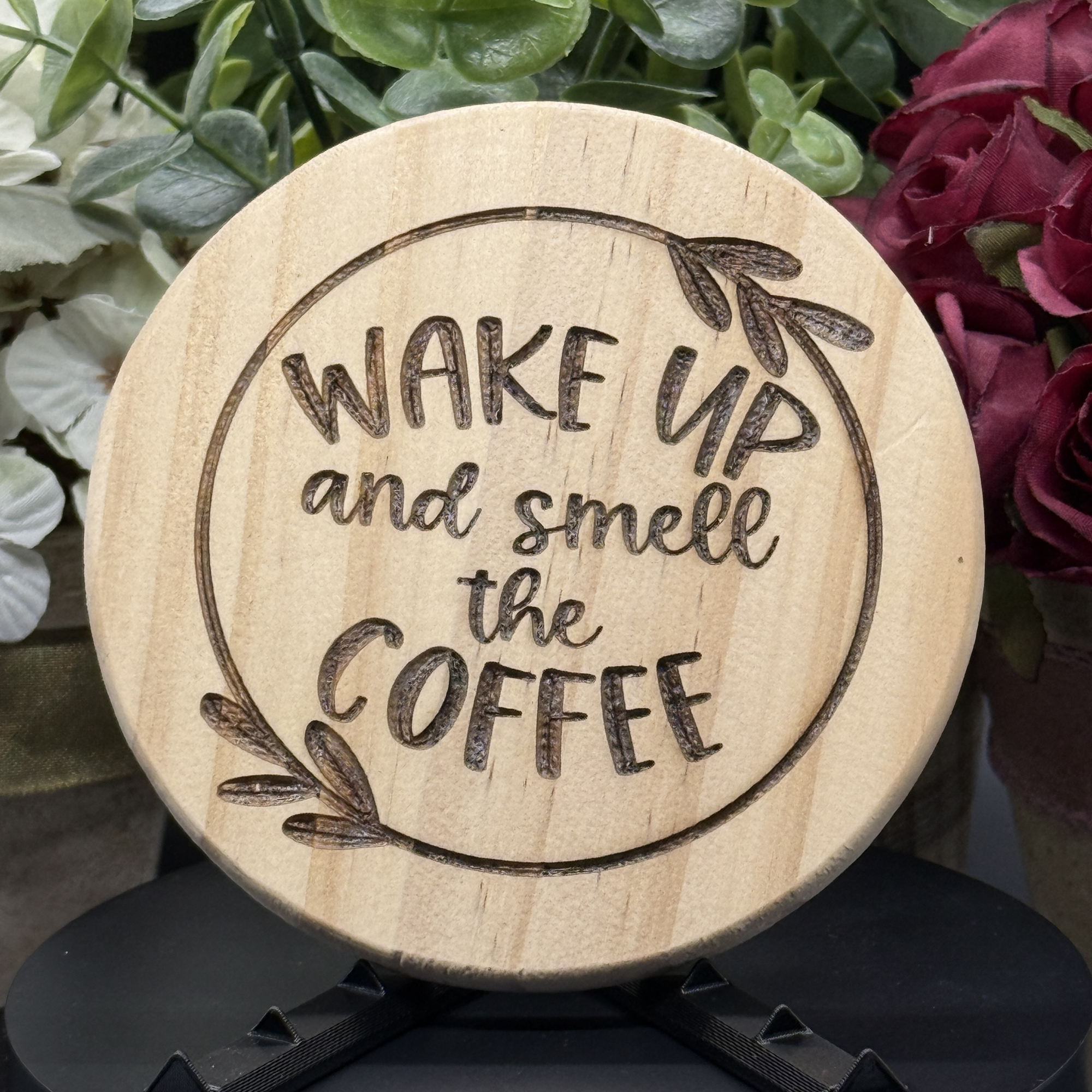 Round pine wood coaster with engraved phrase ‘Wake Up and Smell the Coffee’ inside a leaf-decorated circle, displayed with floral background