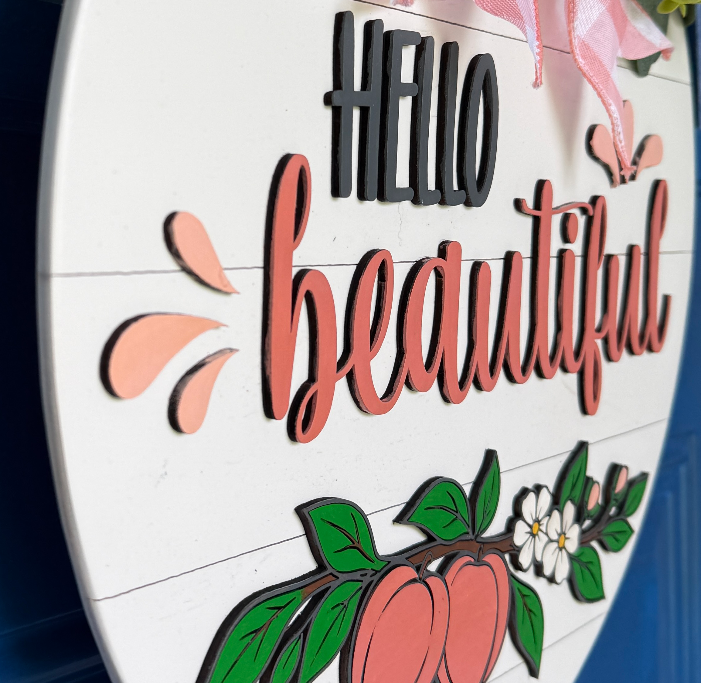 Angled close-up showing dimensional layered wood construction, painted peach artwork, and script lettering on a peach door hanger.