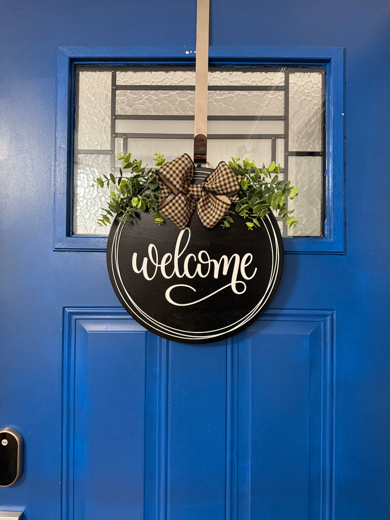 Full front door display featuring black welcome door hanger with greenery accents and buffalo plaid ribbon bow