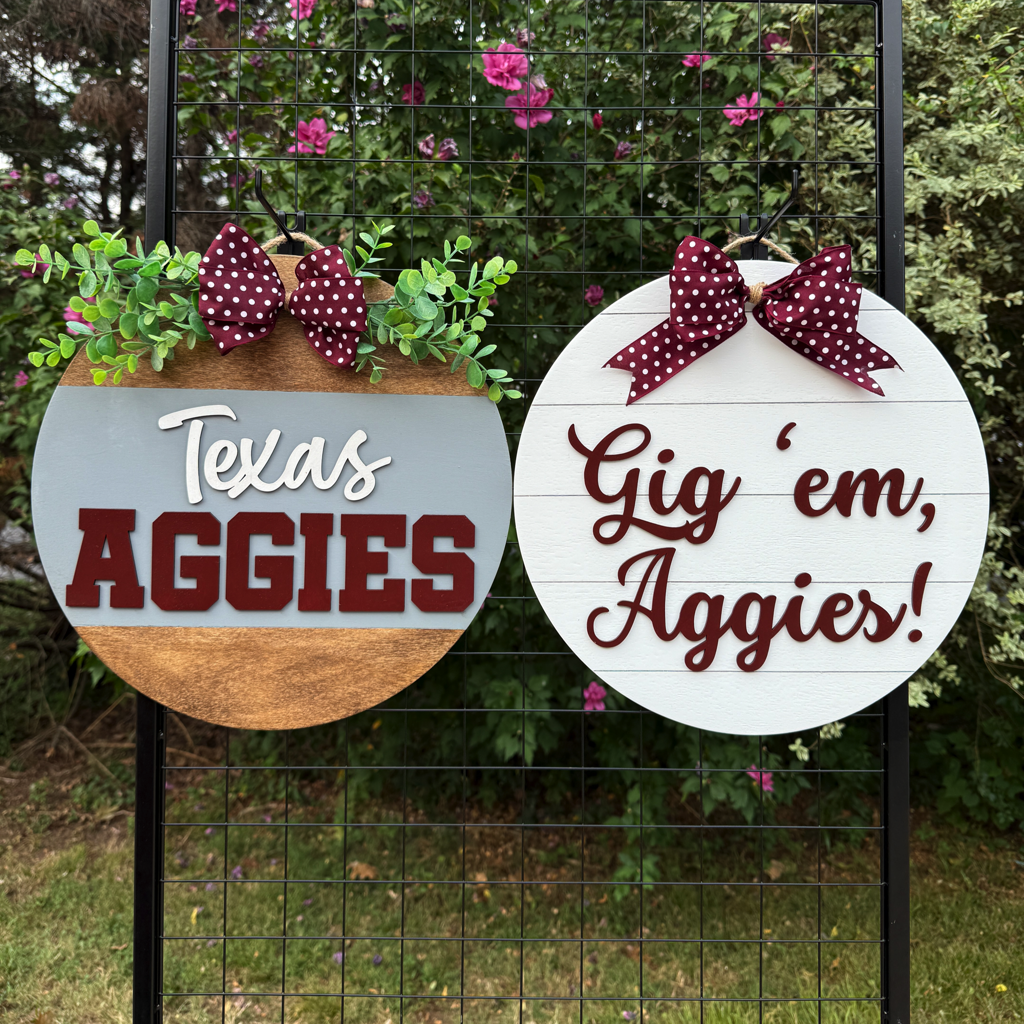 Two round Texas A&M-themed door hangers displayed outdoors on a black grid with greenery in the background; one reads “Texas Aggies” and the other “Gig ’em, Aggies!”