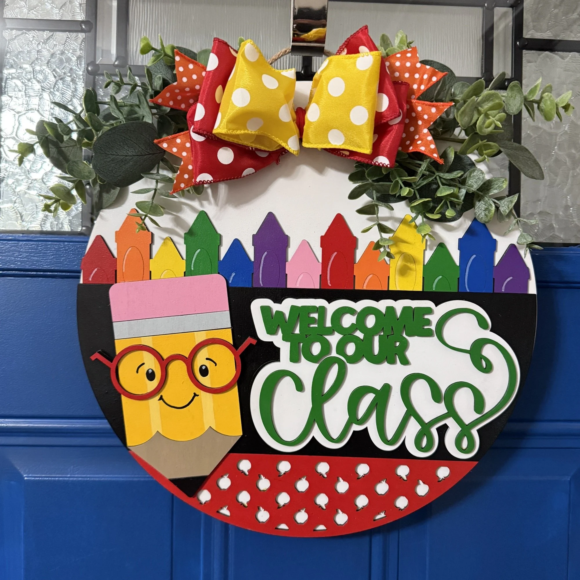 Daytime view of “Welcome to Our Class” door hanger featuring smiling pencil, rainbow crayon row, and red/yellow polka dot bow with greenery on a dark blue door.