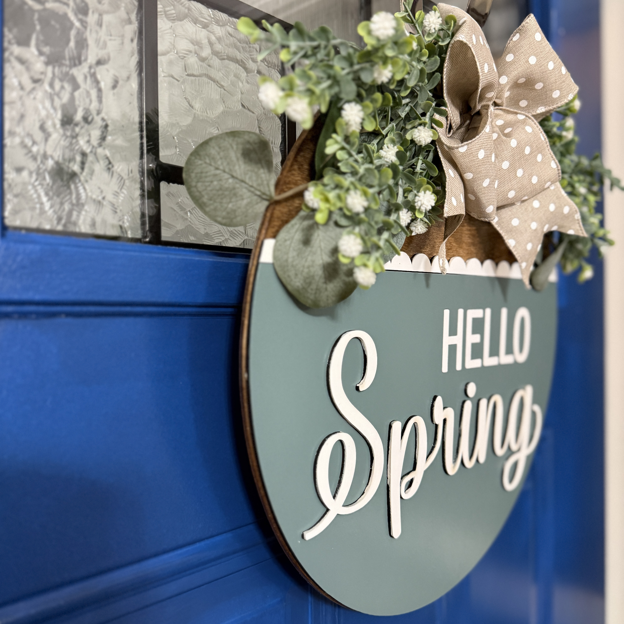 Angled side view of Hello Spring wood door hanger showing layered thickness, greenery detail, and dimensional white script lettering