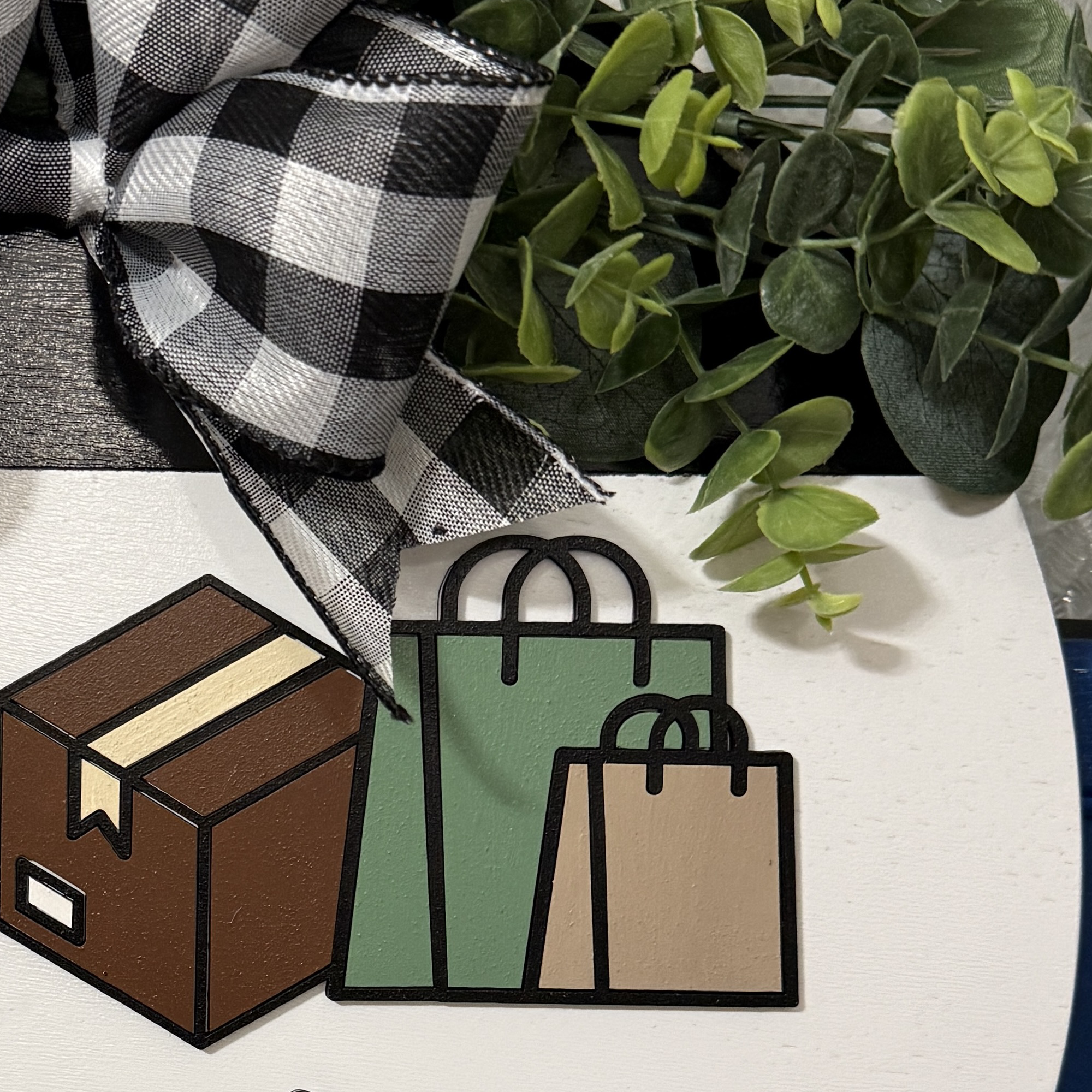 Close-up of layered brown package and green shopping bag graphics on white wooden door hanger with buffalo plaid ribbon and greenery accents.