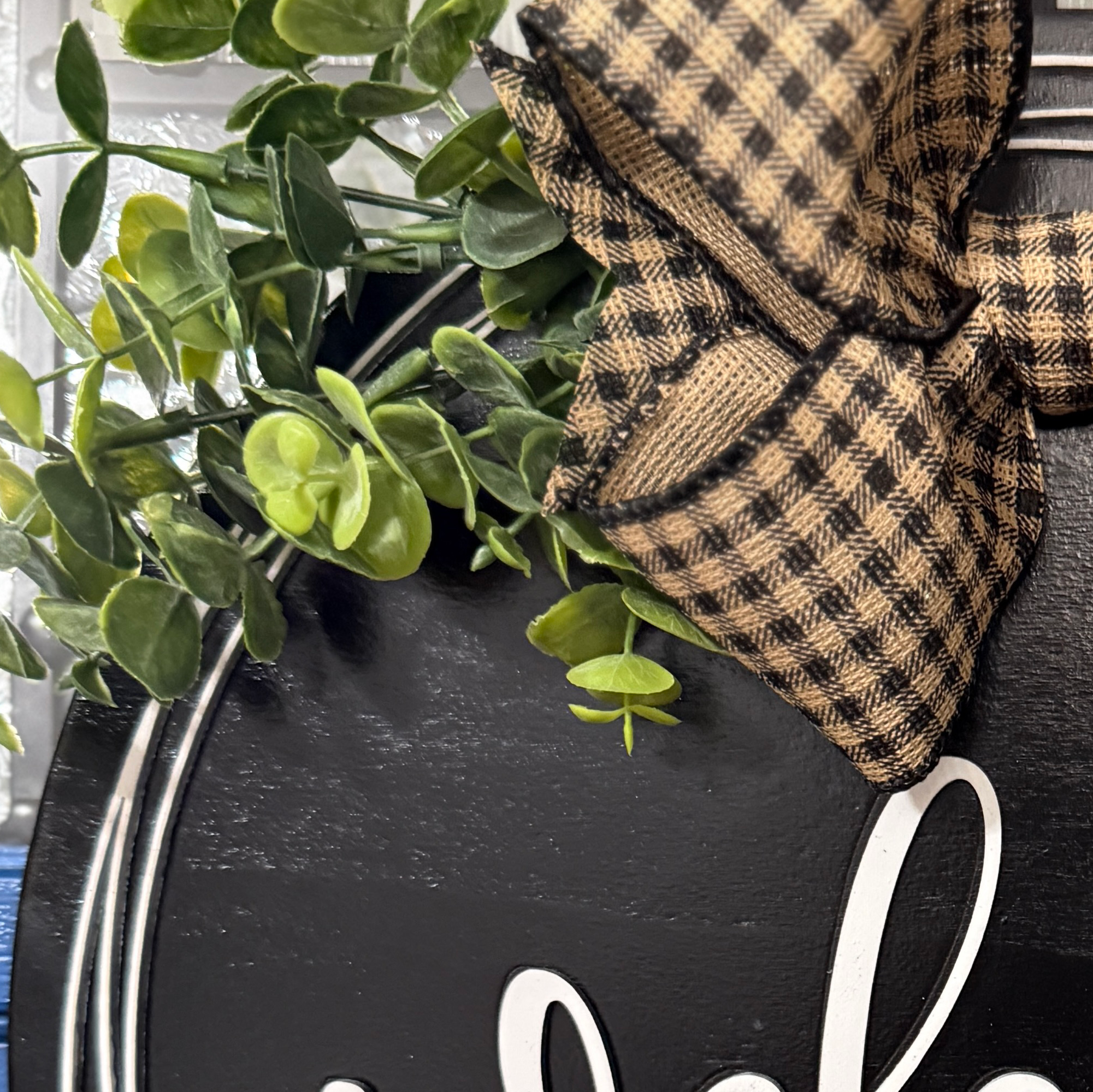 Close-up of faux greenery and tan buffalo plaid bow attached to handcrafted welcome wood door hanger