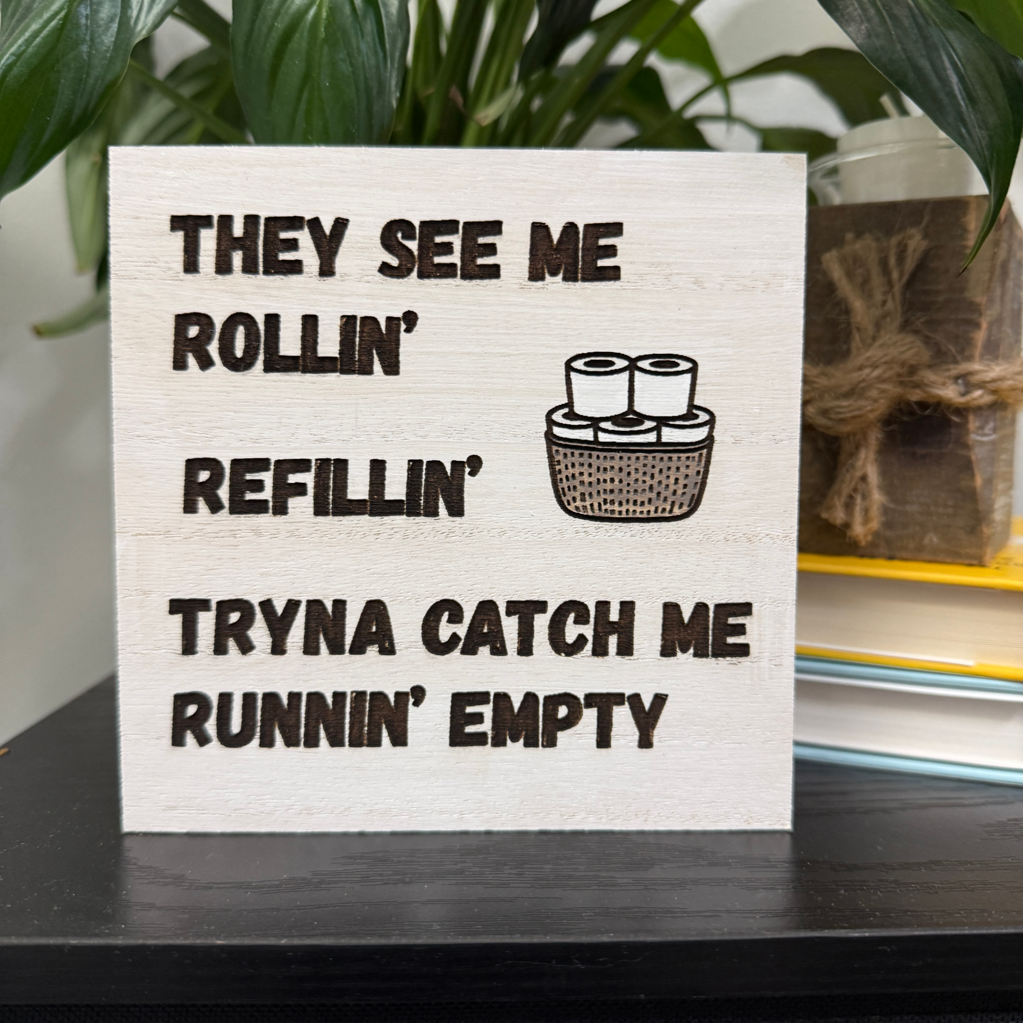 Close-up of humorous toilet paper themed sign with engraved text and roll illustration on white-washed wood, styled beside greenery and stacked books