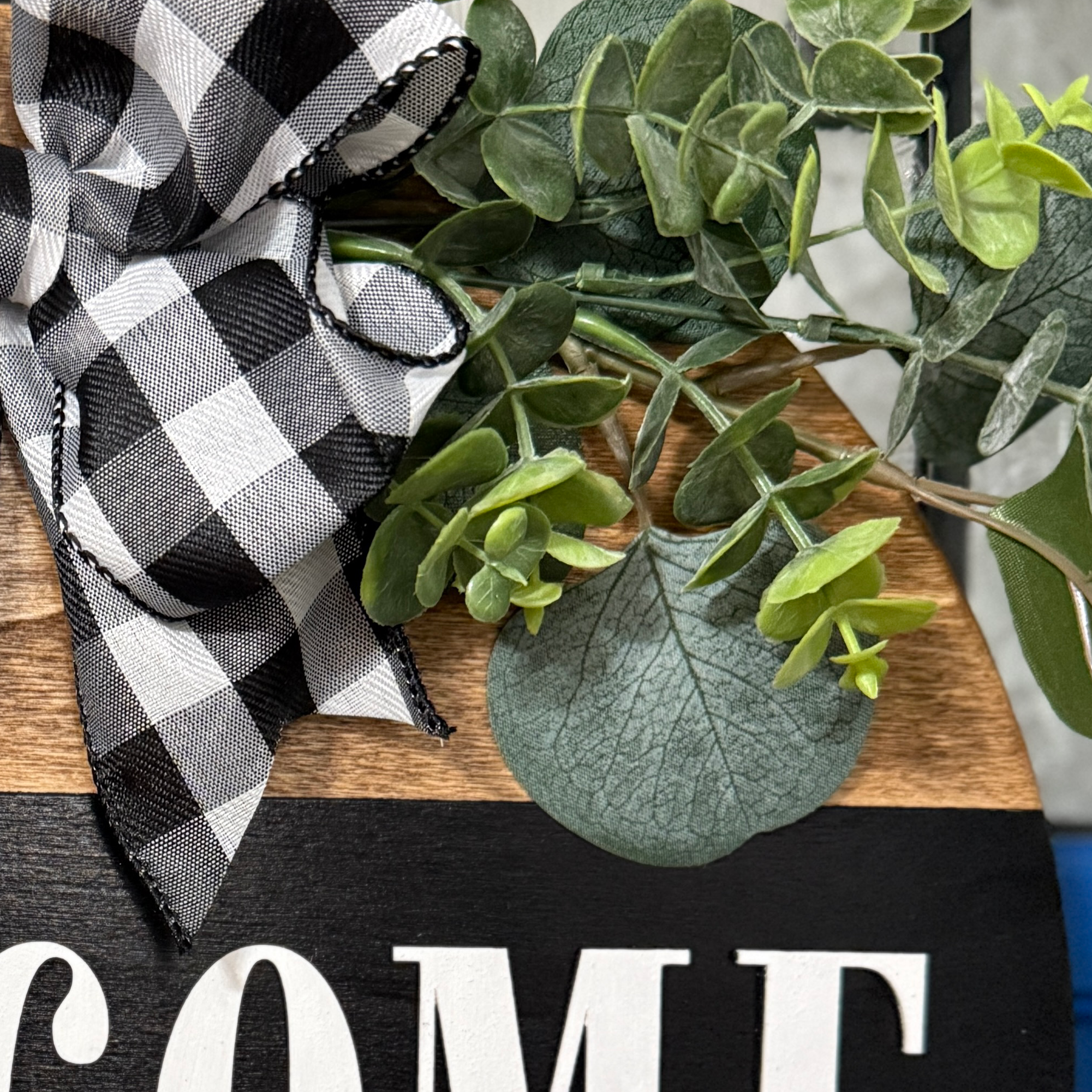 Detailed view of buffalo plaid ribbon and greenery accents on rustic welcome to our home wood hanger