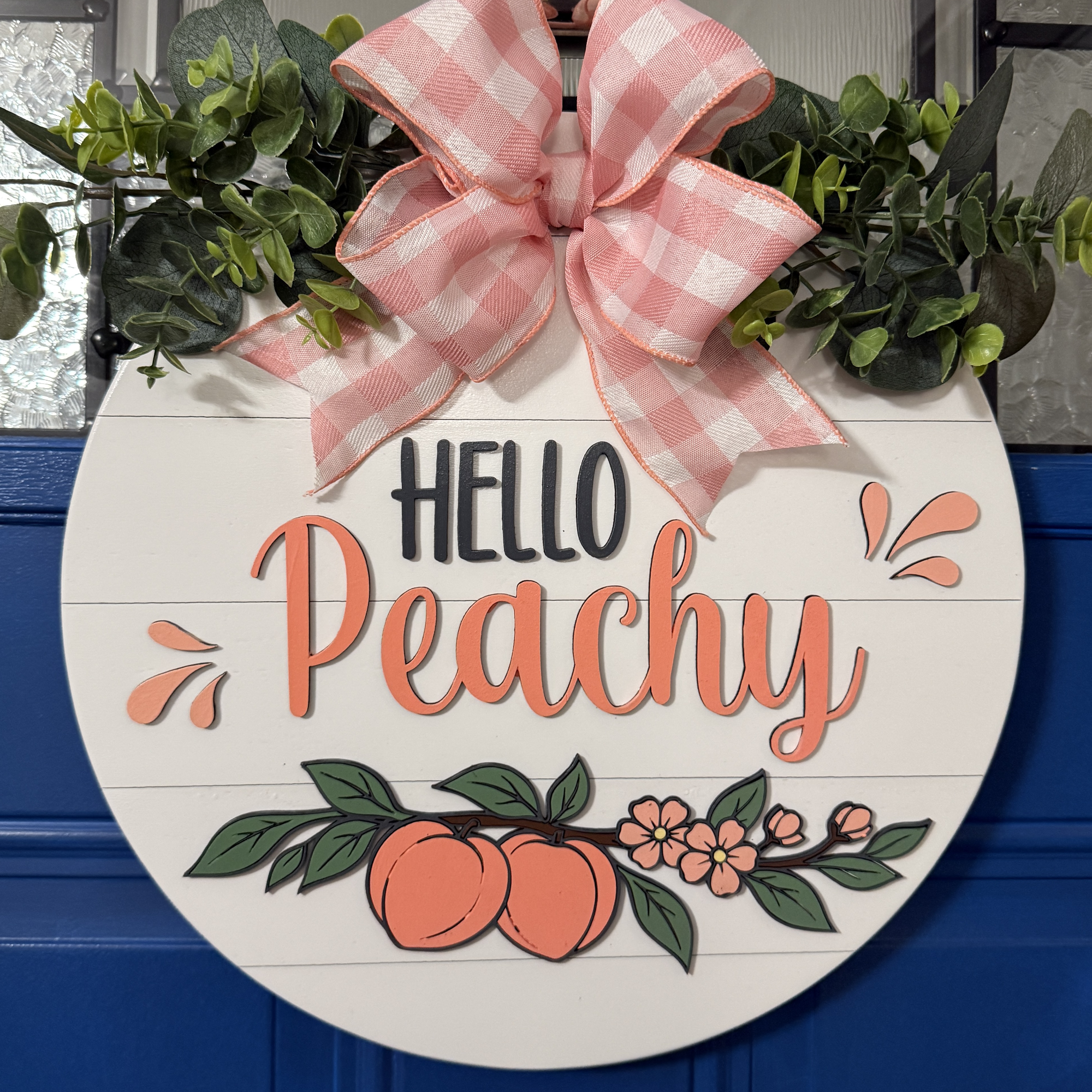 Front view of Hello Peachy round wood door hanger with raised peach lettering, hand-painted peaches, and pink gingham bow on blue door