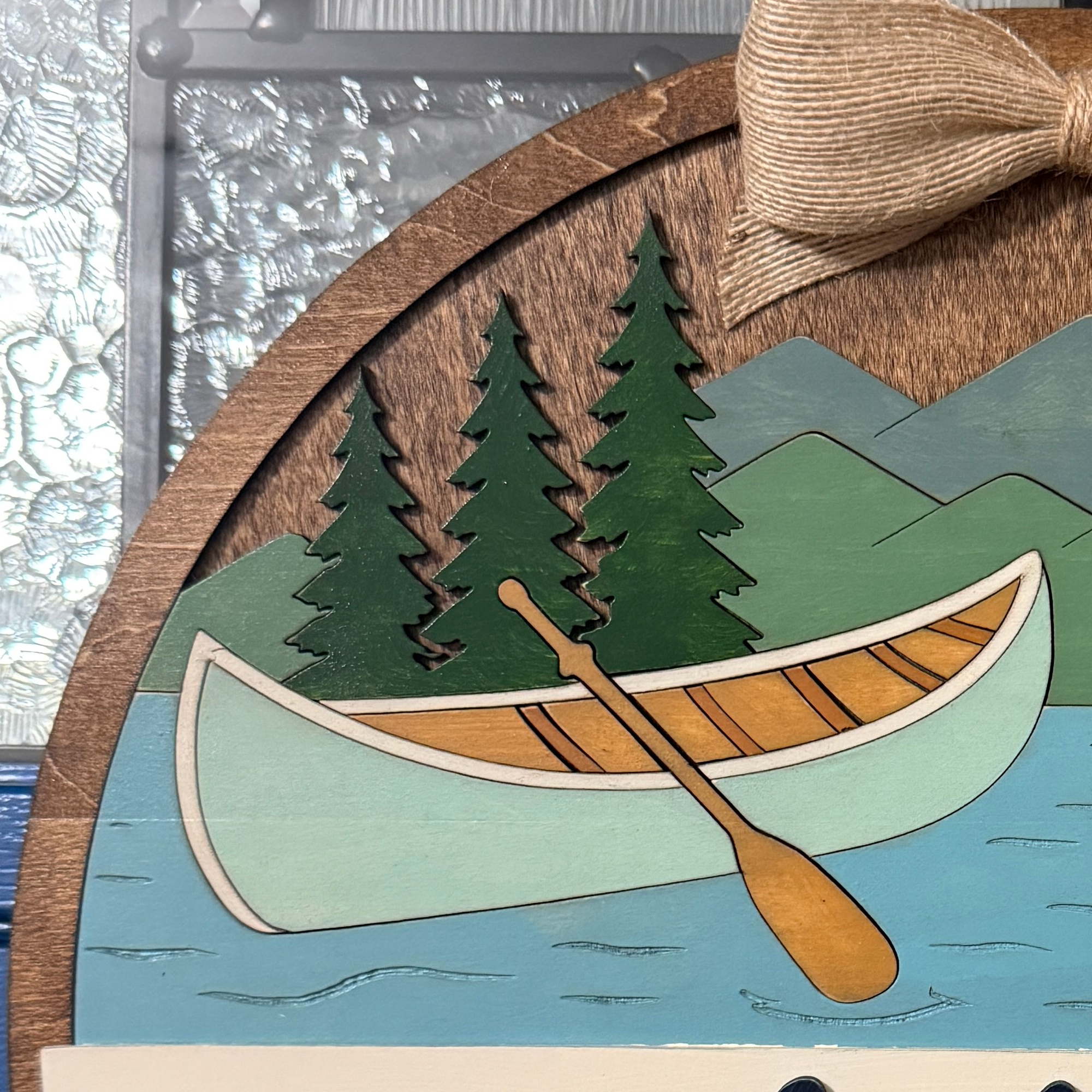 Close-up of canoe, pine trees, mountains, and lake scene on the layered wood sign.