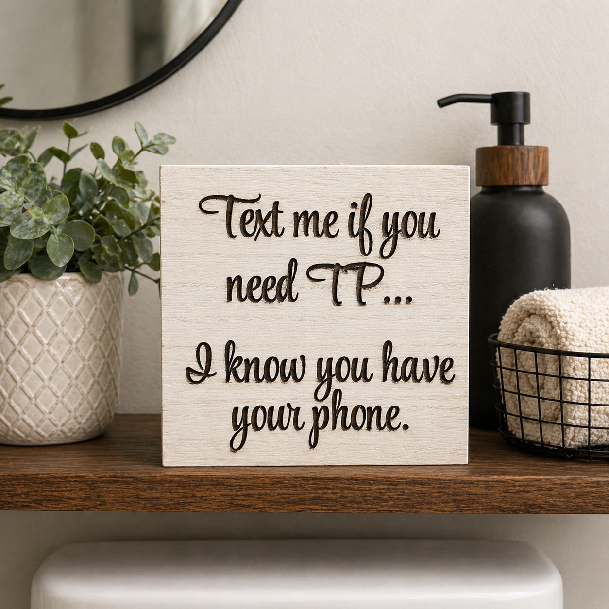 Bathroom shelf sitter sign with engraved “Text me if you need TP… I know you have your phone” displayed on a wooden shelf beside soap dispenser and greenery