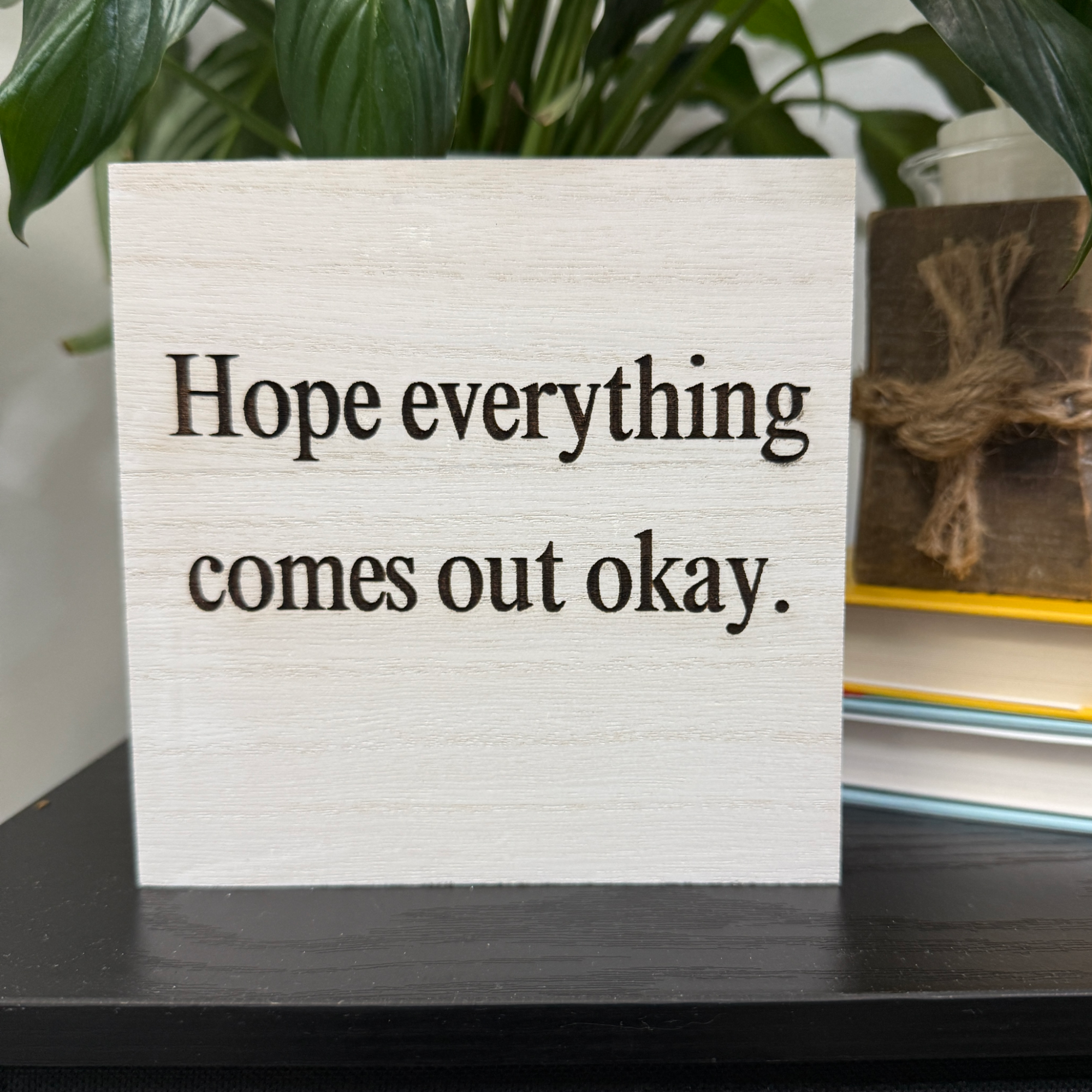 Close-up of white-washed wood sign with engraved “Hope everything comes out okay” text, styled beside greenery and stacked books