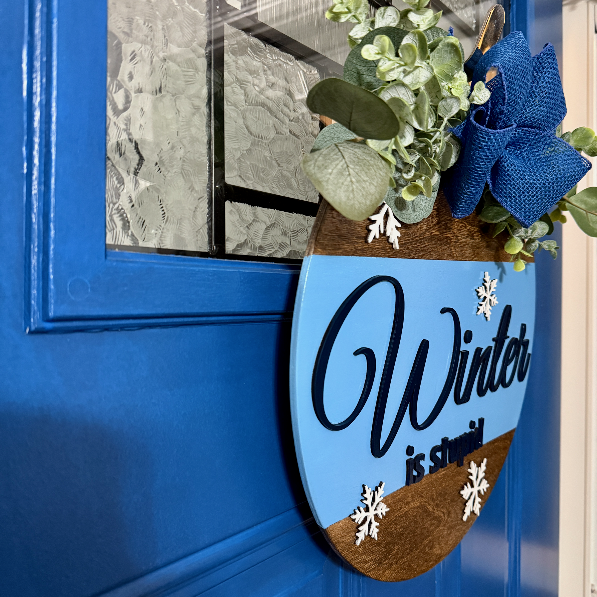 Angled side view of Winter Is Stupid winter door hanger showing layered wood depth and jute rope hanging on a blue door