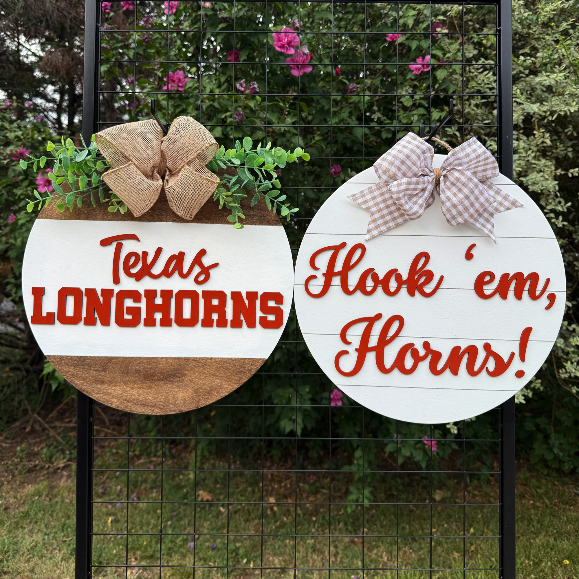 Two round Texas-themed door hangers displayed outdoors on a black grid with greenery in the background; one reads “Texas Longhorns” and the other “Hook ’em, Horns!”