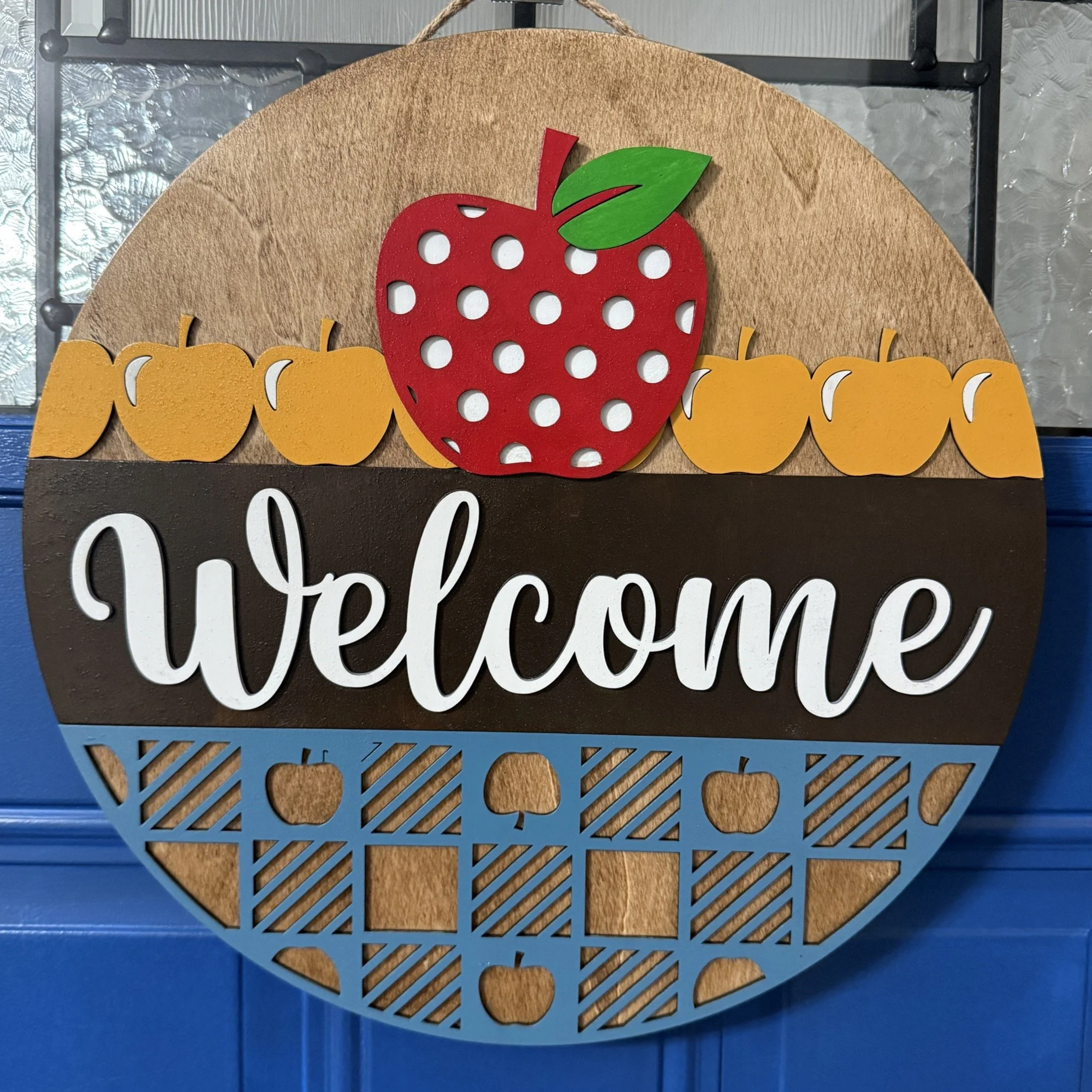 14-inch layered wood “Welcome” door hanger with red polka-dot apple, golden apples, and blue patterned base with apple cutouts on a blue front door.