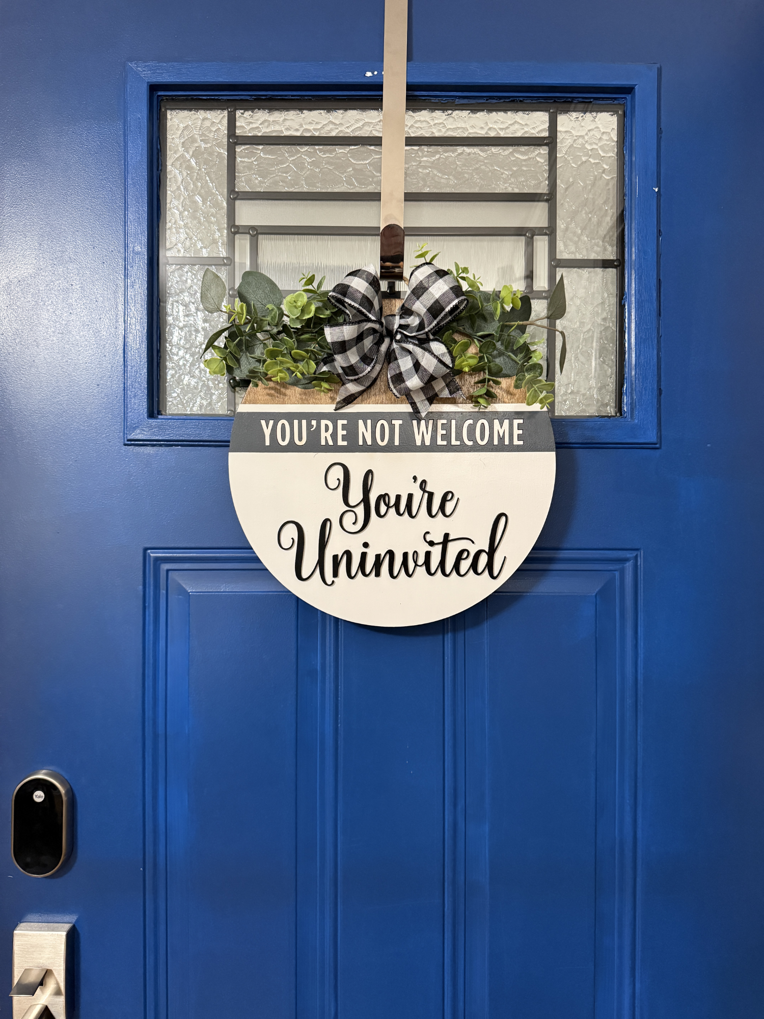 Full view of blue front door with round farmhouse-style wooden door hanger featuring “You’re Not Welcome, You’re Uninvited” phrase, greenery, and buffalo plaid bow.