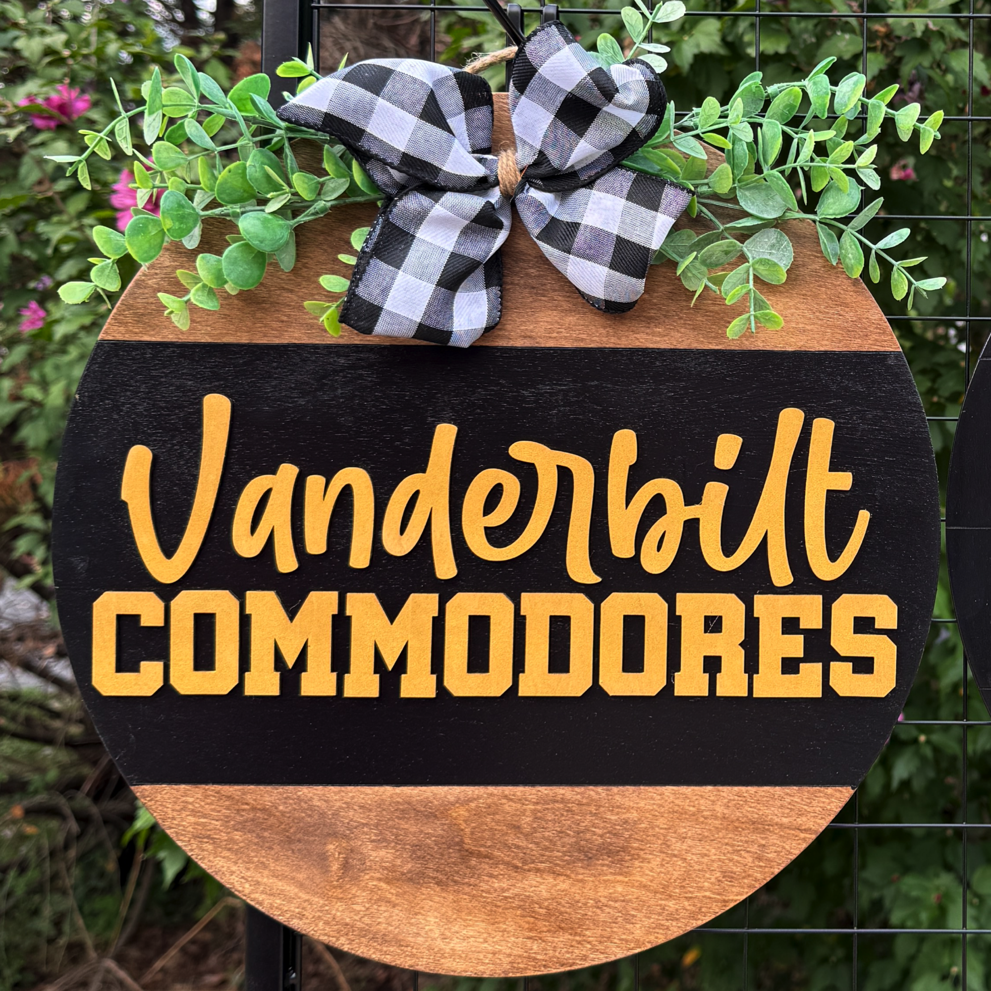 Round Vanderbilt Commodores door hanger with gold lettering, greenery accents, and buffalo plaid bow on rustic wood background