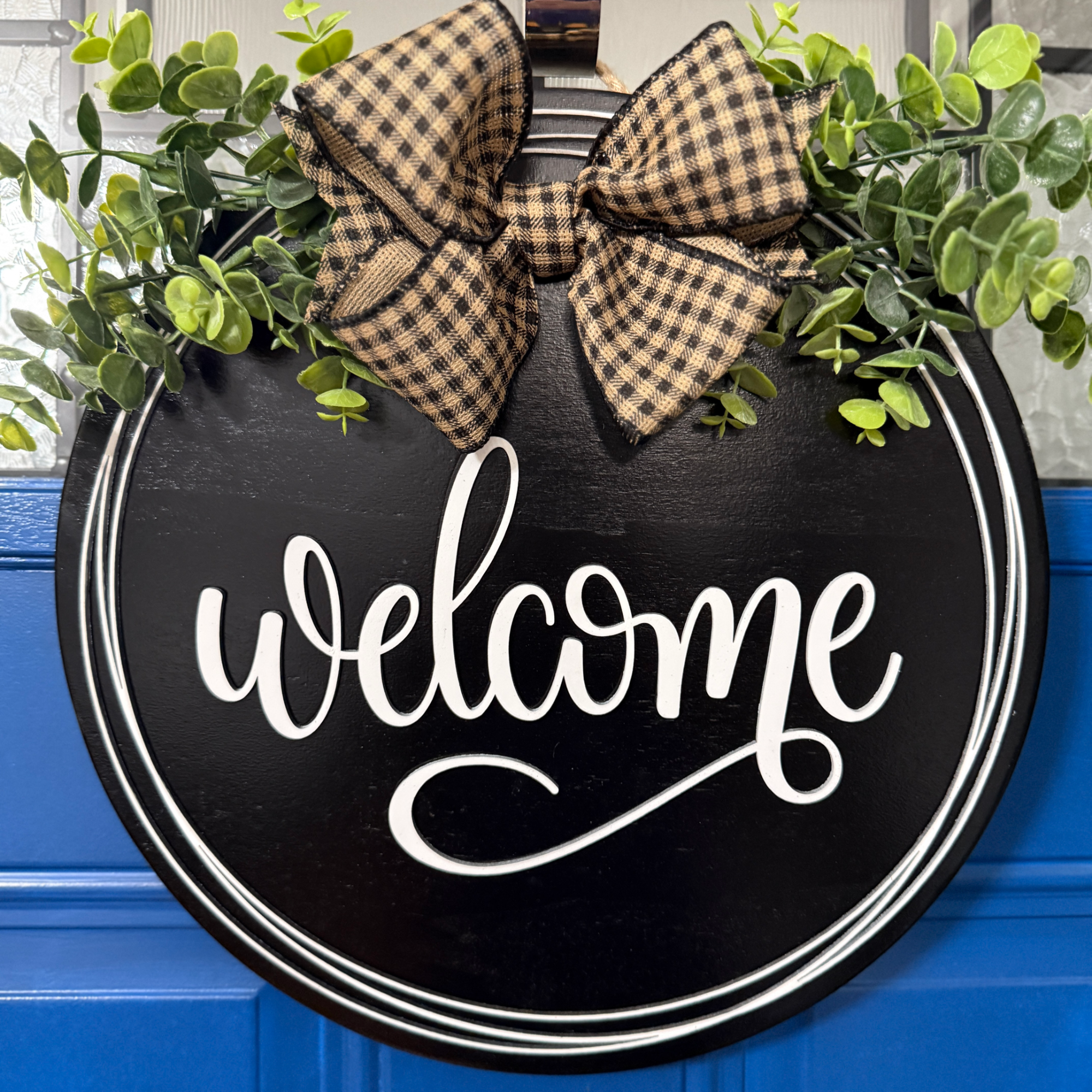 Black round welcome door hanger with white script lettering, greenery accents, and tan buffalo plaid bow on blue front door