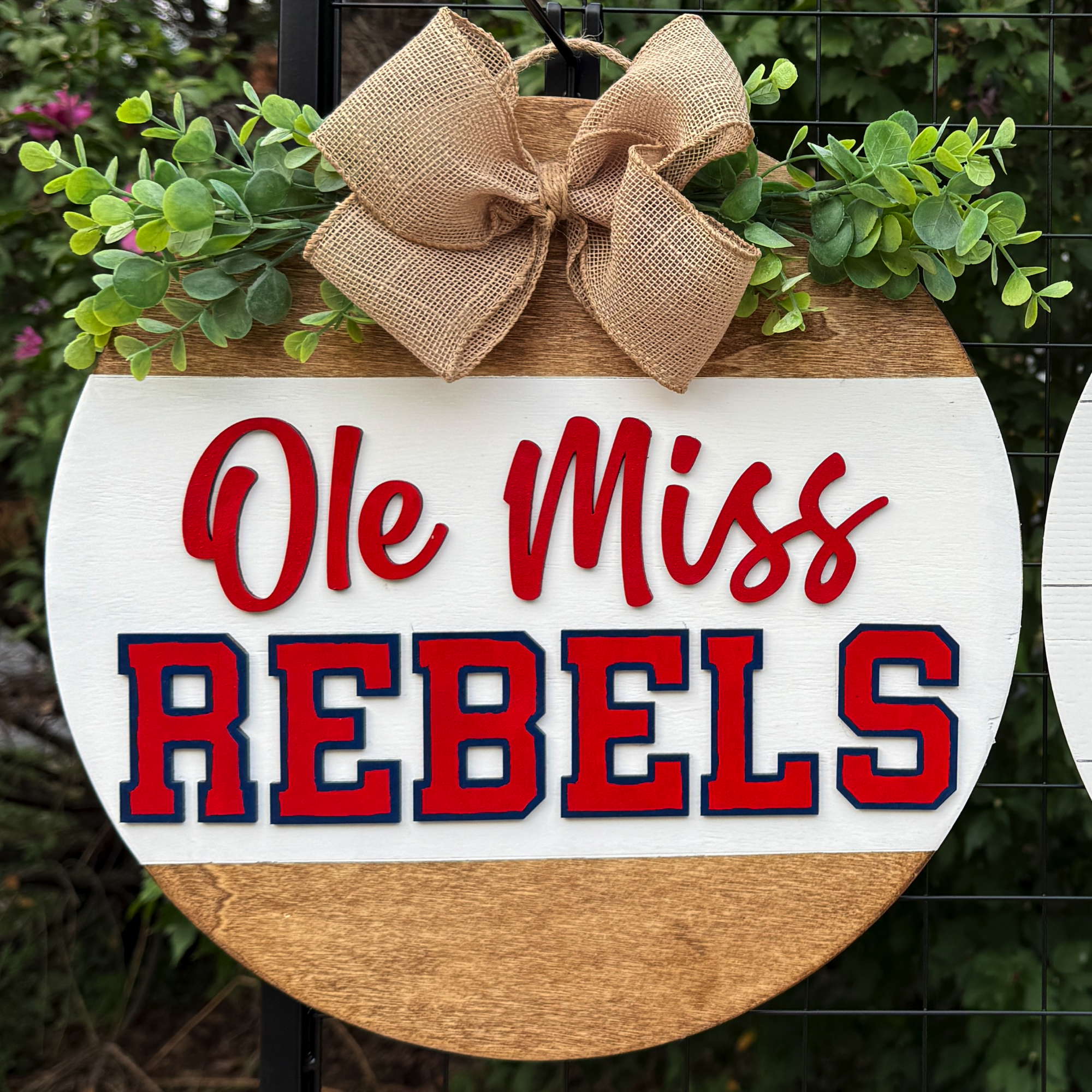 Round Ole Miss Rebels door hanger with red and blue lettering, greenery accents, and burlap bow on rustic wood background