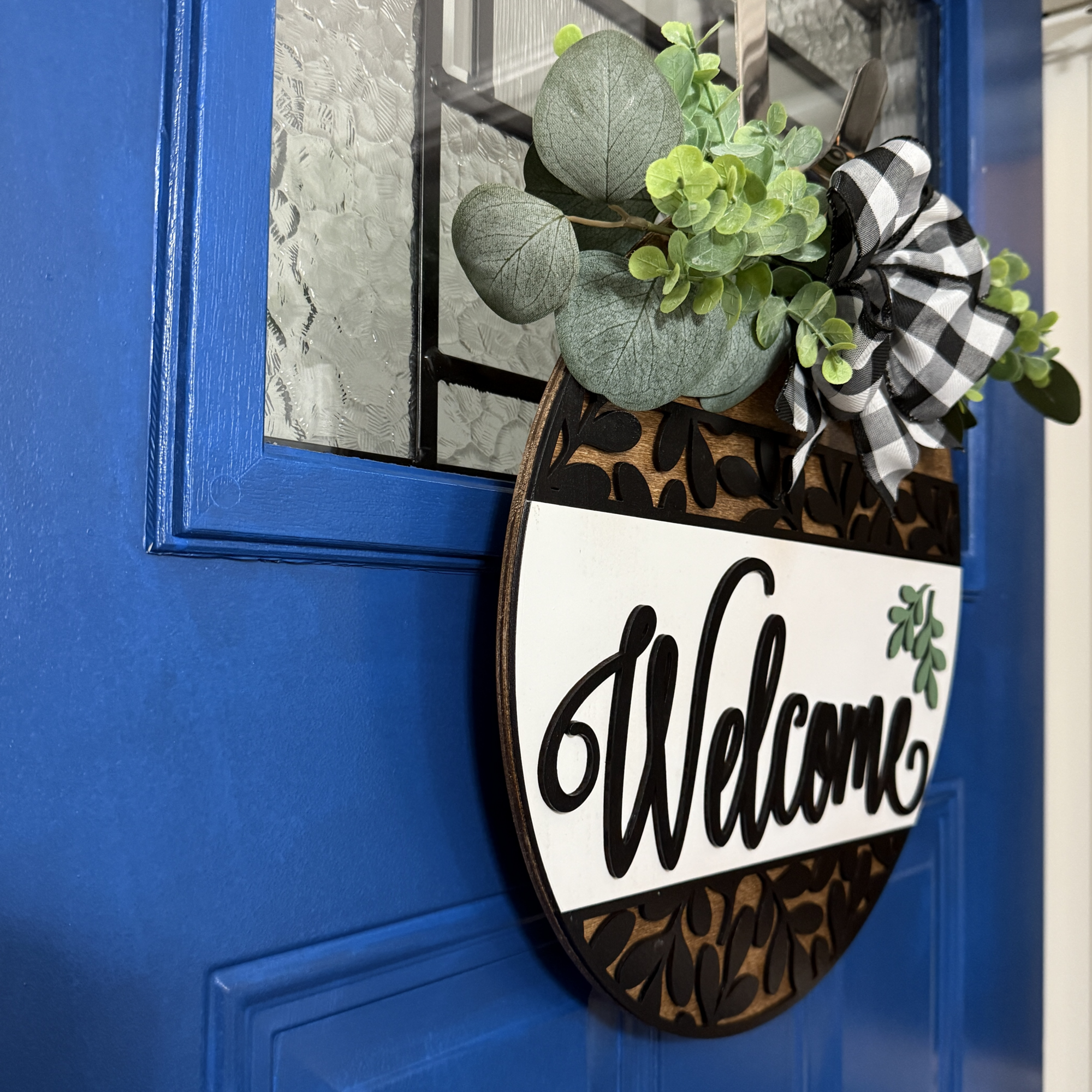 Side view of round buffalo plaid welcome door hanger with greenery, layered leaf design, and white center panel