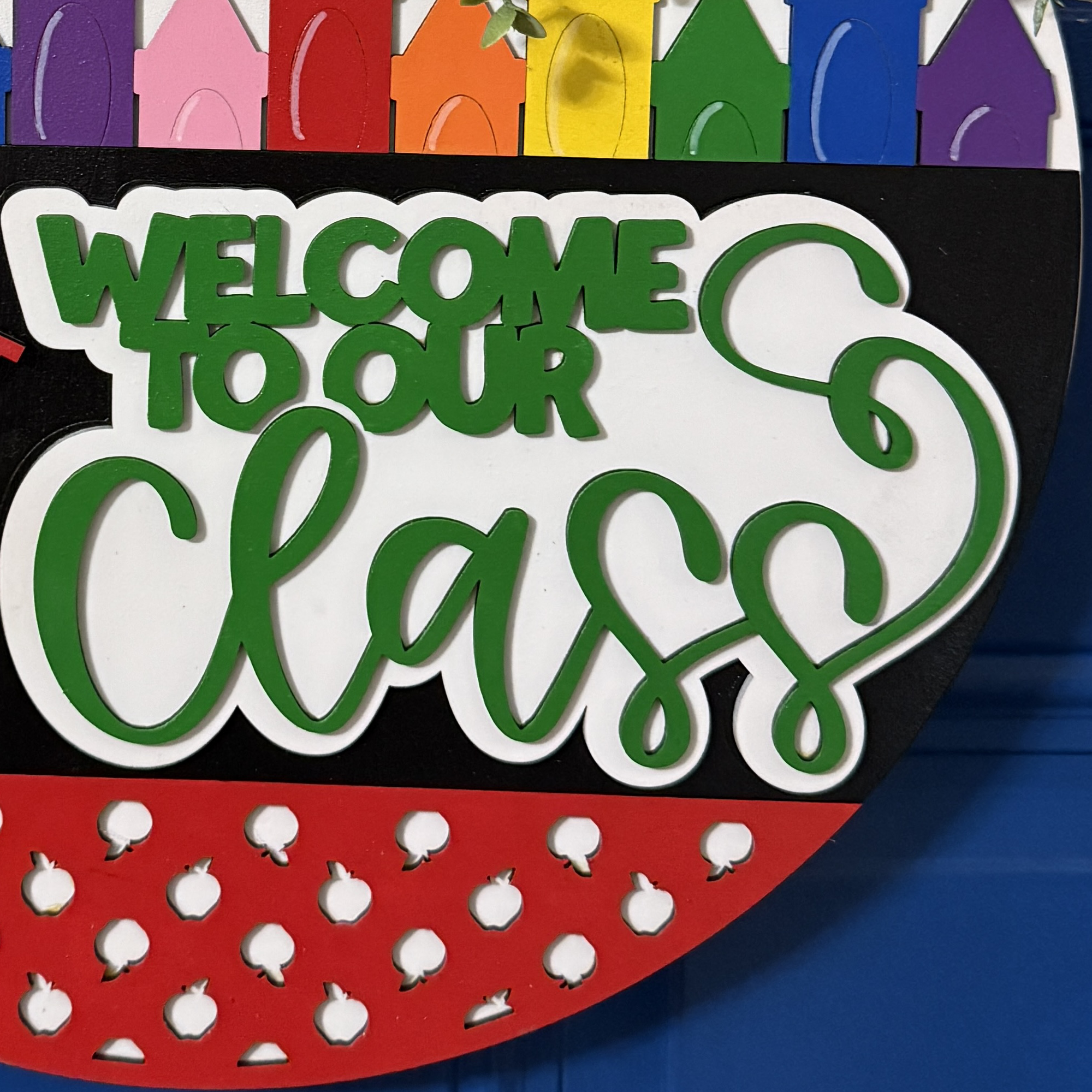Side view of “Welcome to Our Class” door hanger featuring smiling pencil, rainbow crayon row, and red/yellow polka dot bow with greenery on a dark blue door.