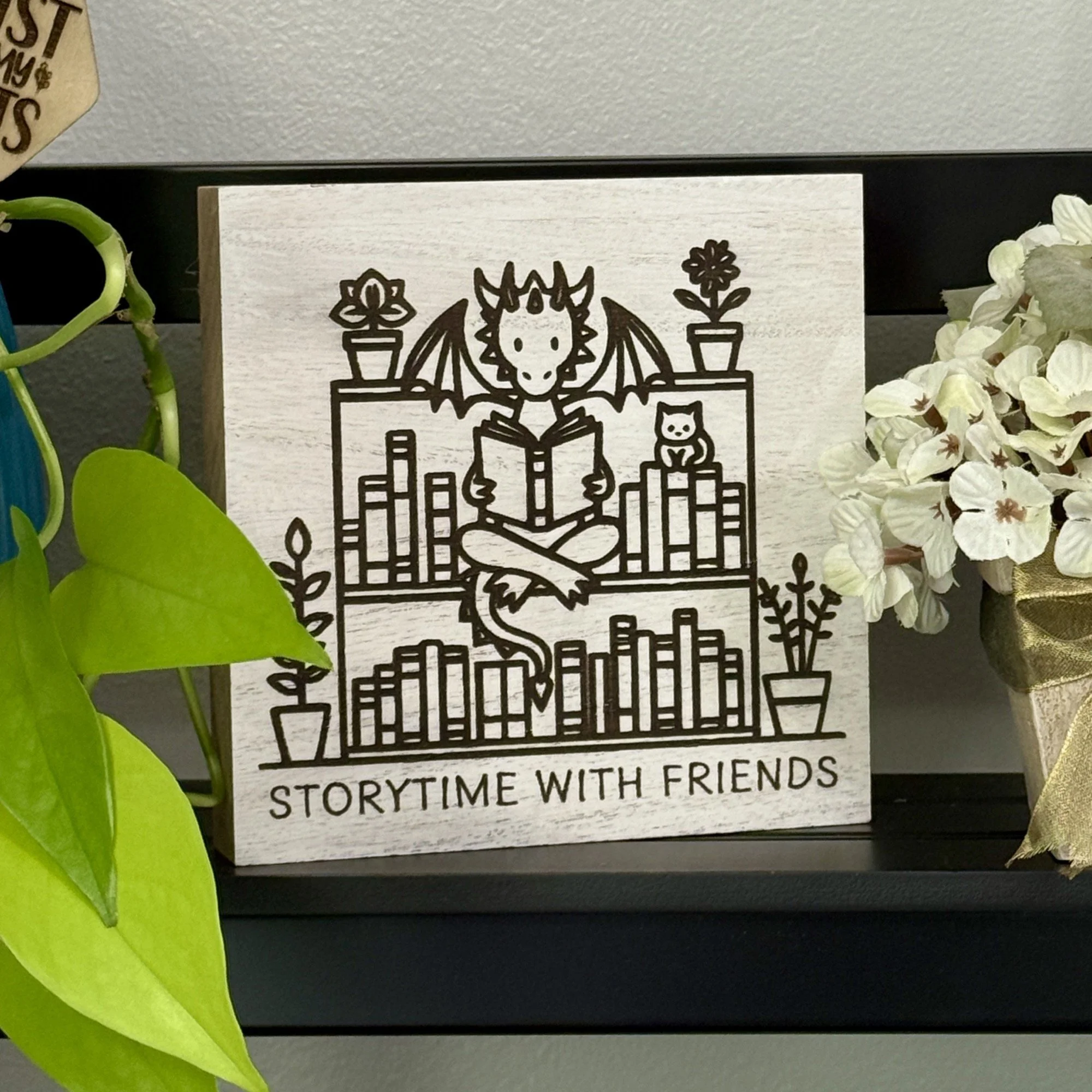 White-washed wood shelf sitter engraved with a dragon reading a book beside a small cat, framed by potted plants on a tall bookshelf, displayed on a black shelf with surrounding decor.