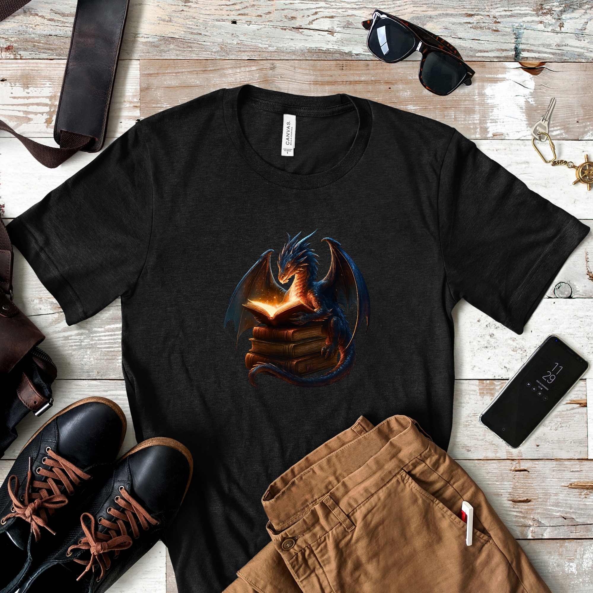 Flat lay of heathered black t-shirt with glowing book dragon design surrounded by boots, jeans, and accessories