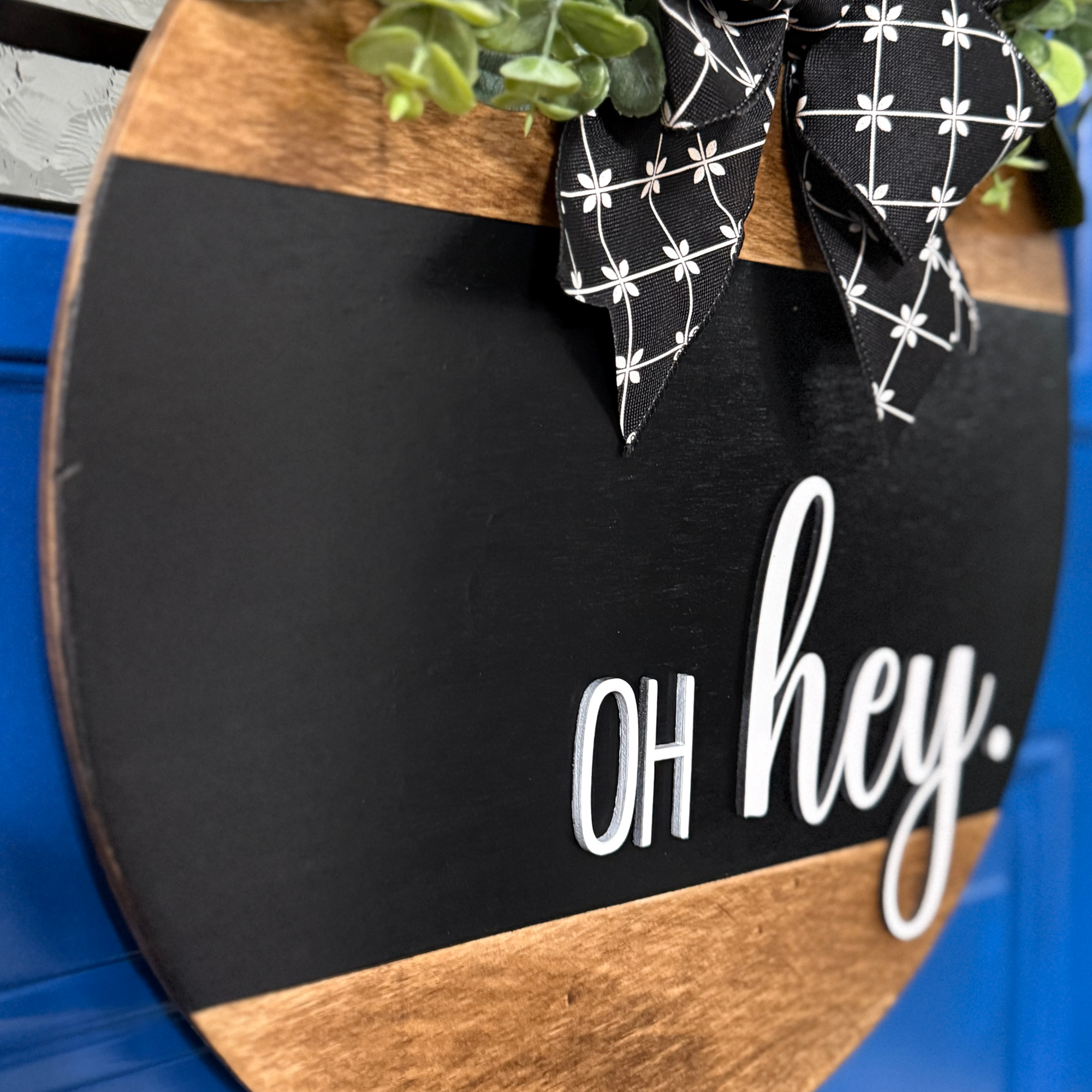 Side angle view of black and natural wood oh hey door hanger with layered wood detail and decorative bow